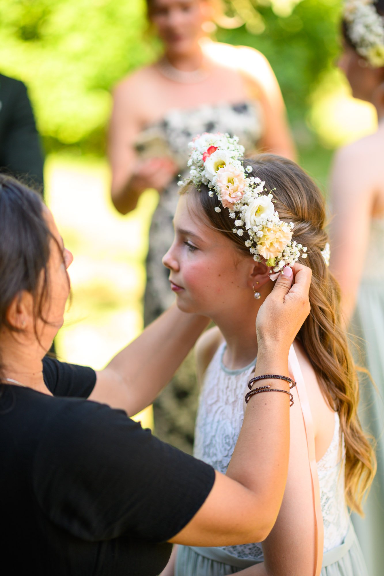 Une jeune femme porte une couronne de fleurs et reçoit de l'aide pour la mettre à son cheveu lors d'une cérémonie en plein air.