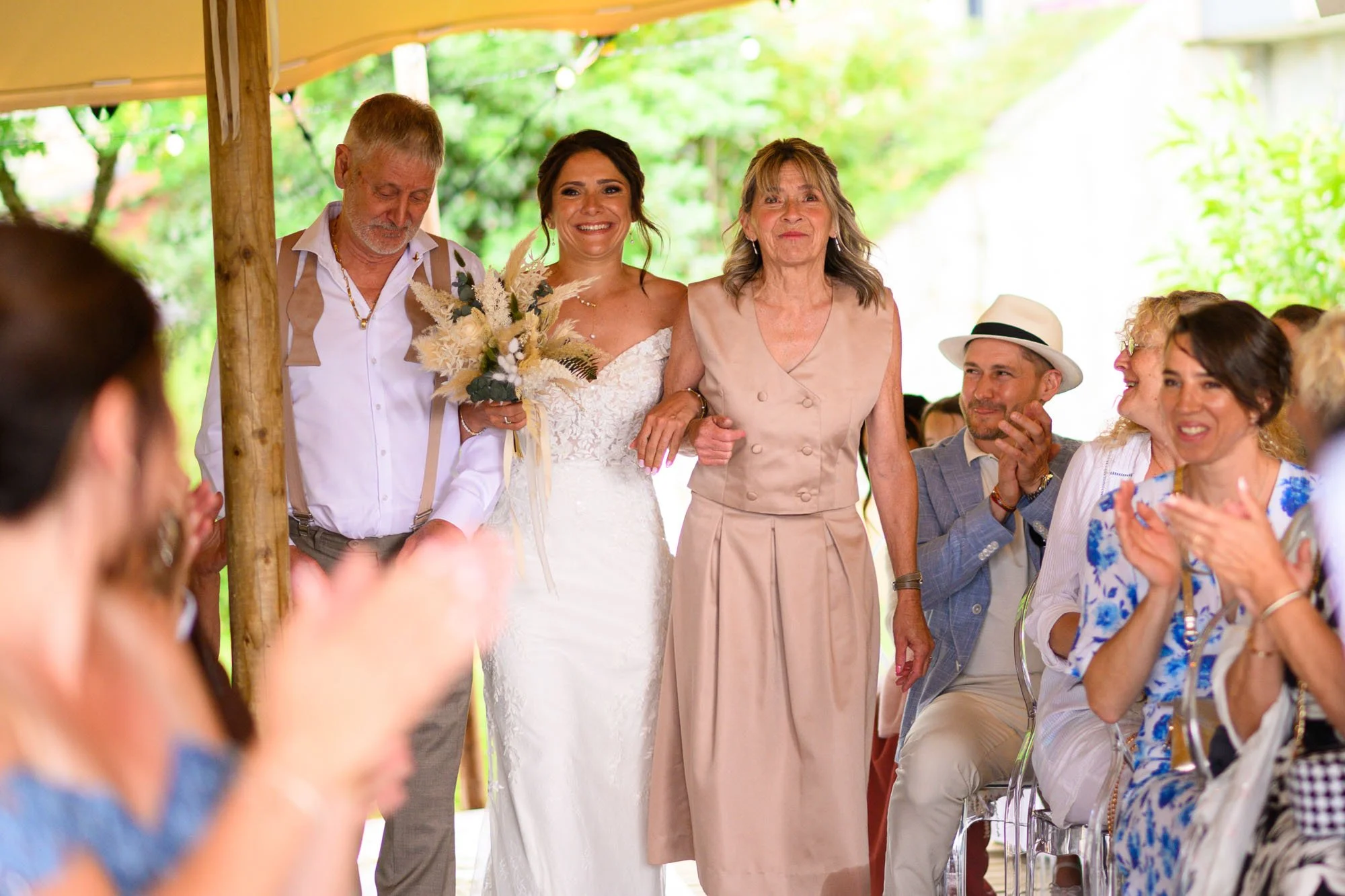Une mariée en robe blanche est aidée à marcher par une femme âgée lors d'un mariage en plein air, entourée d'invités applaudissant et souriant