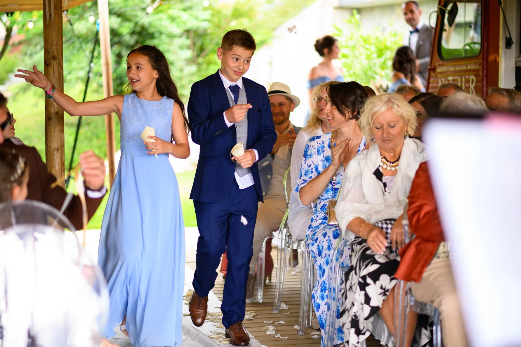 Des enfants en costume de mariage marchent dans une cérémonie en plein air, entourés de convives assis. La scène est colorée, avec un décor naturel en arrière-plan.