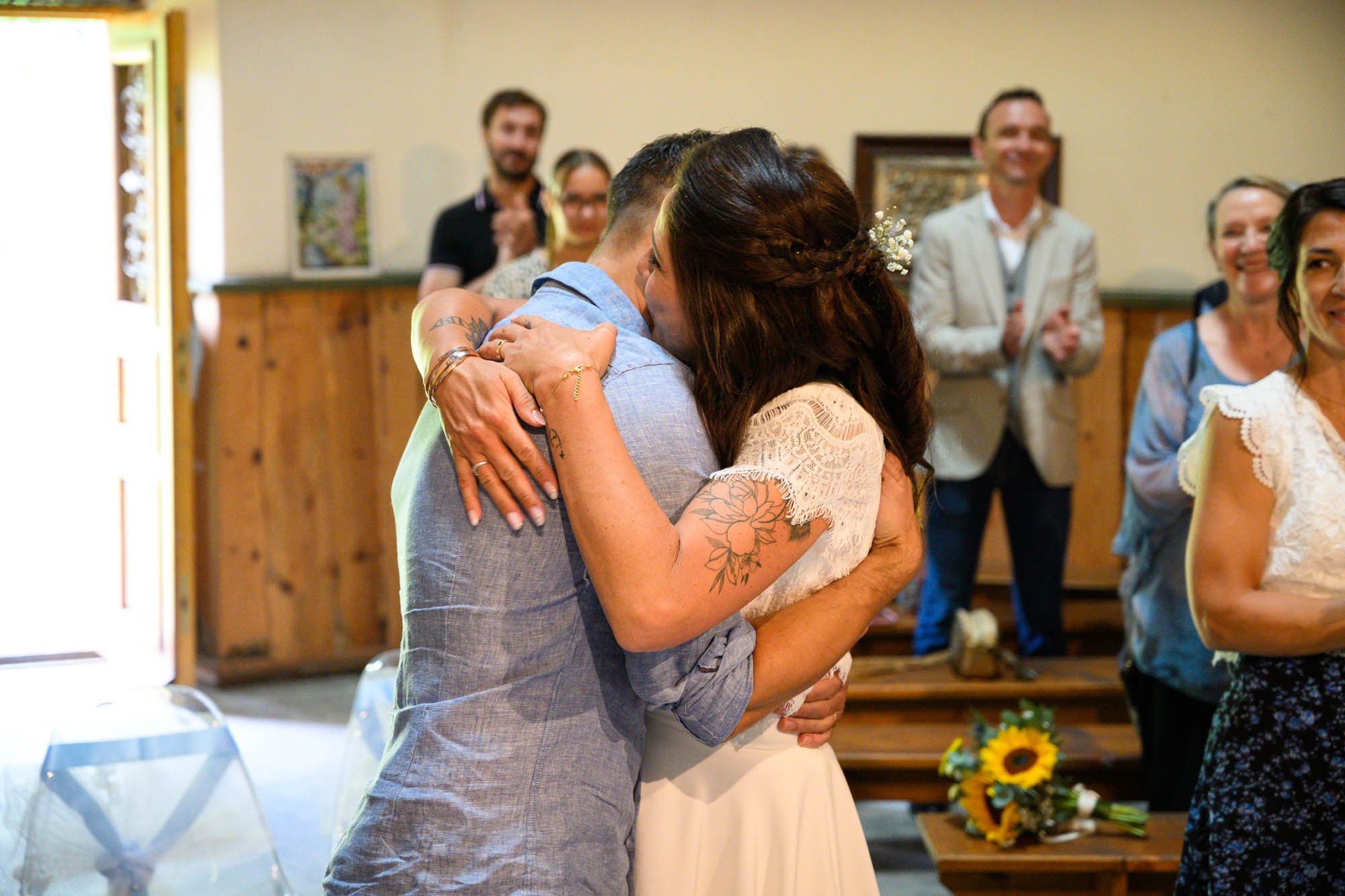 Un couple s'embrasse lors d'une cérémonie ou d'une réception, entouré de plusieurs personnes souriantes.