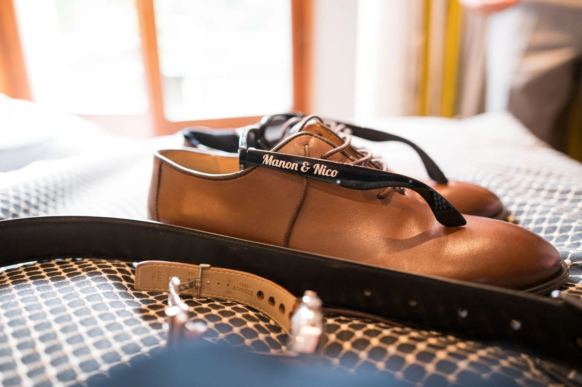 Une paire de chaussures en cuir marron avec des lacets noirs portant l'inscription 'Manon & Nico' reposent sur une surface. En premier plan, un ceinture noire et une autre beige sont visibles.