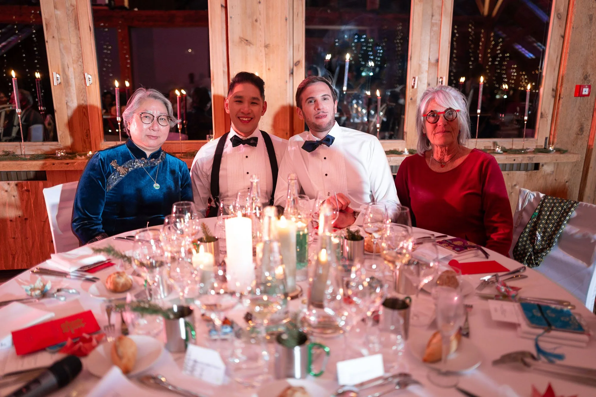 Un groupe de quatre personnes assises autour d'une table décorée pour un dîner dans une ambiance chaleureuse avec des bougies, à l'arrière-plan des fenêtres laissant apparaître des lumières de la nuit.