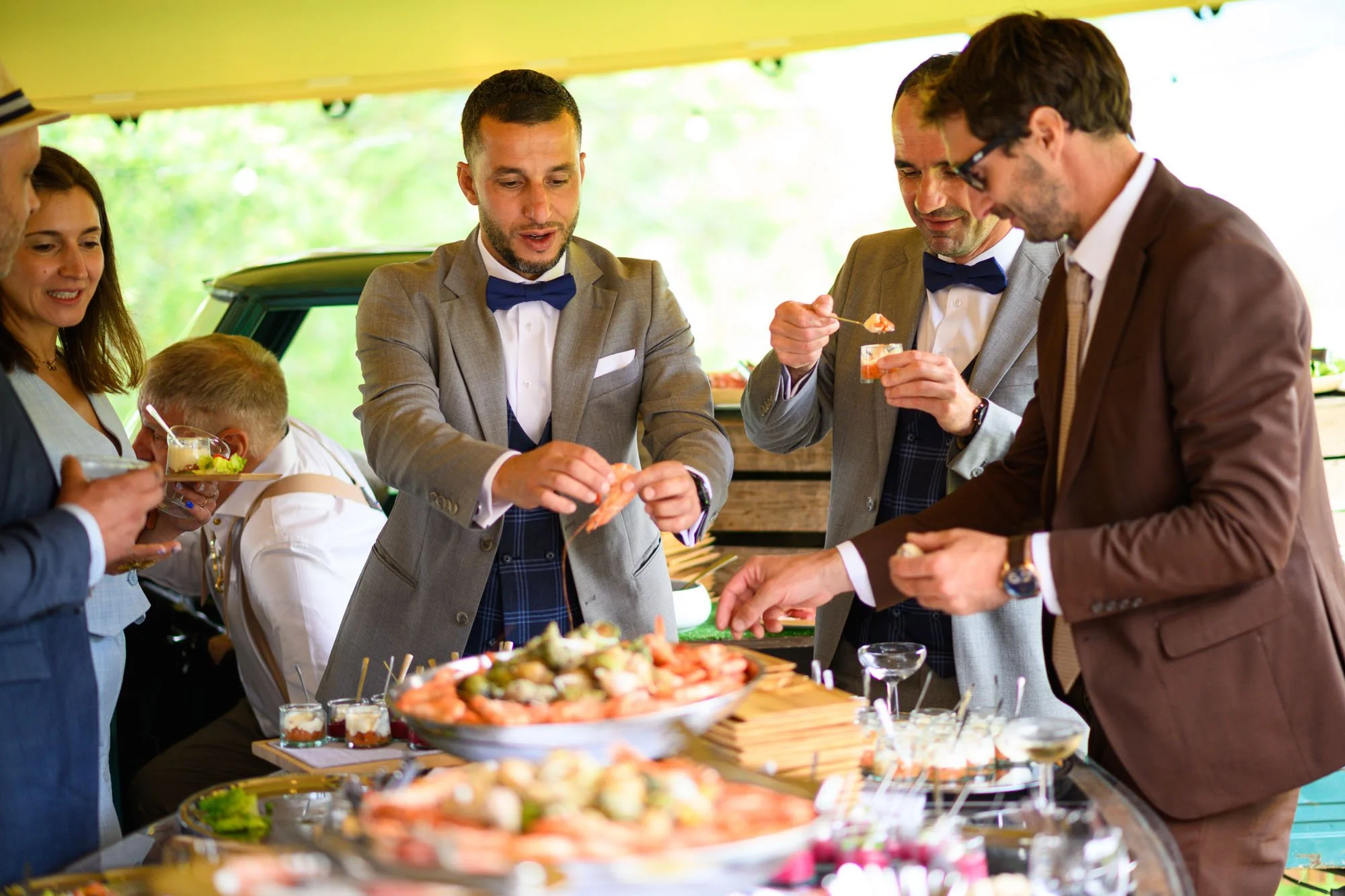 Groupe d'hommes et de femmes en tenues formelles dégustant des plats lors d'une réception en plein air, avec de la nourriture présentée sur une table.