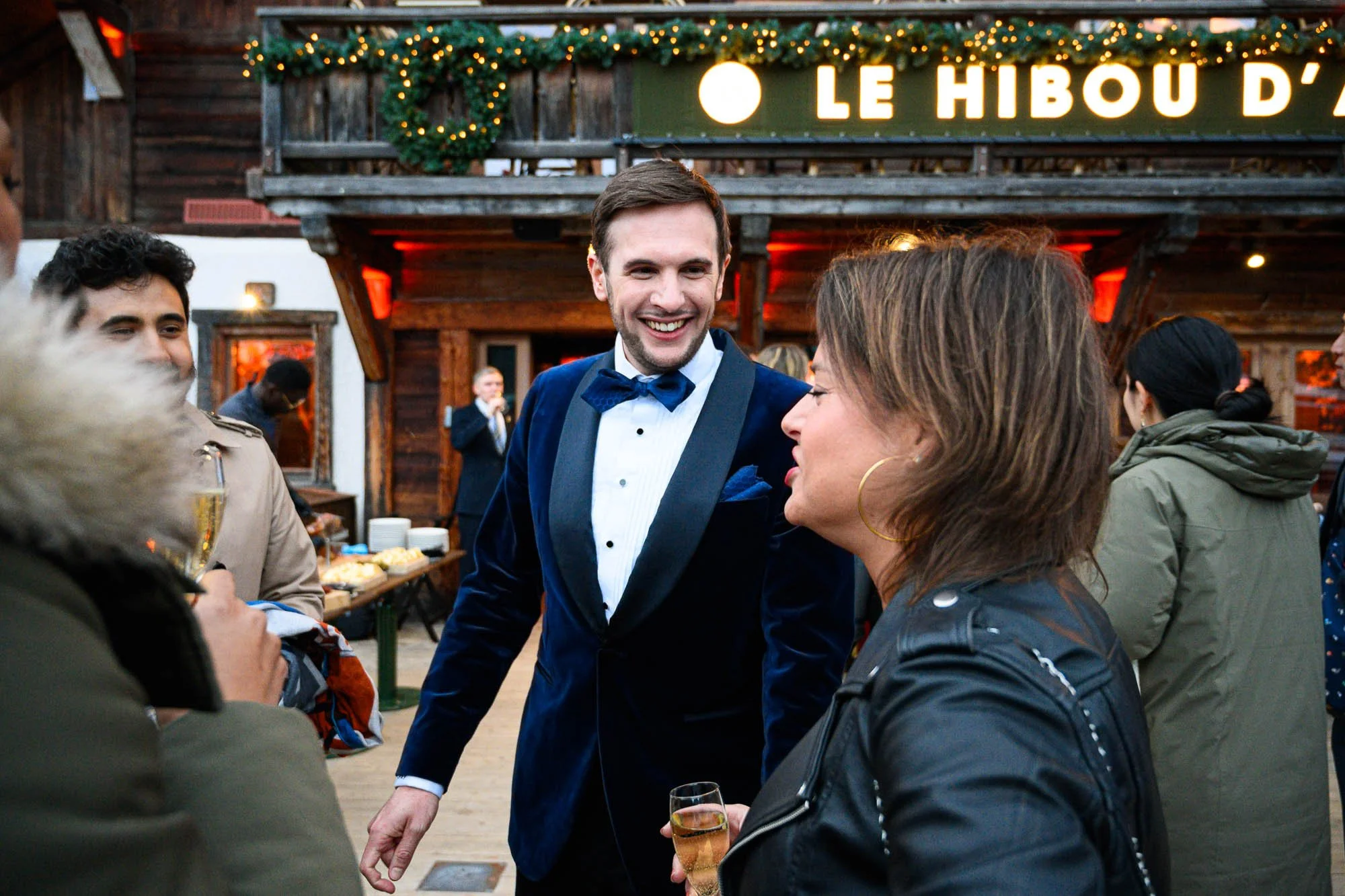 Groupe de personnes lors d'une fête en extérieur, discussions animées, un homme en costume bleu avec une cravate papillon sourit, ambiance festive avec décorations lumineuses.