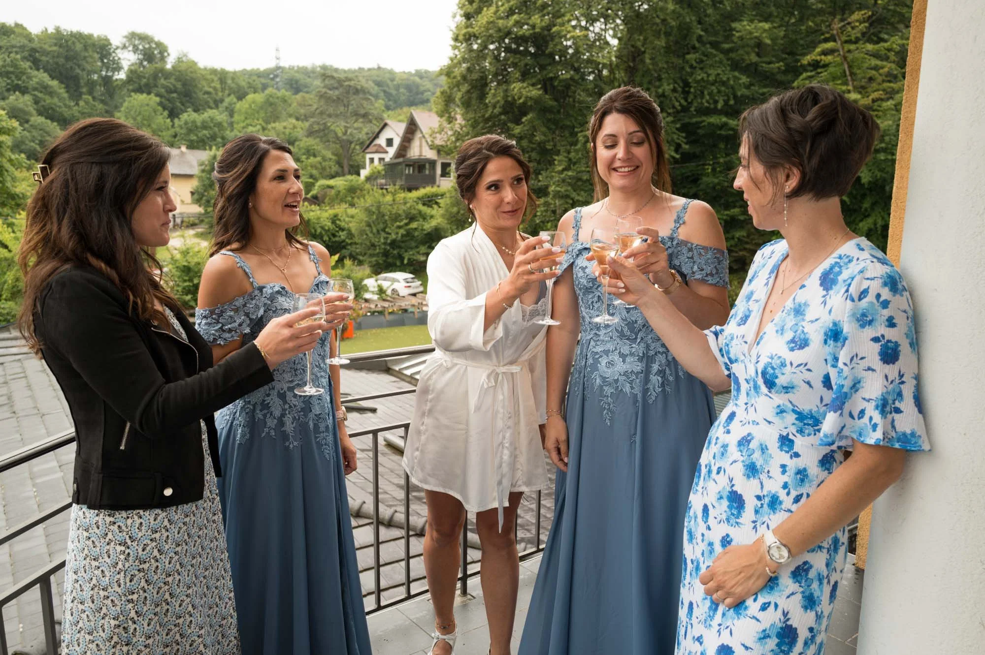 Groupe de cinq femmes faisant un toast sur un balcon, en arrière-plan une nature verdoyante et plusieurs maisons.