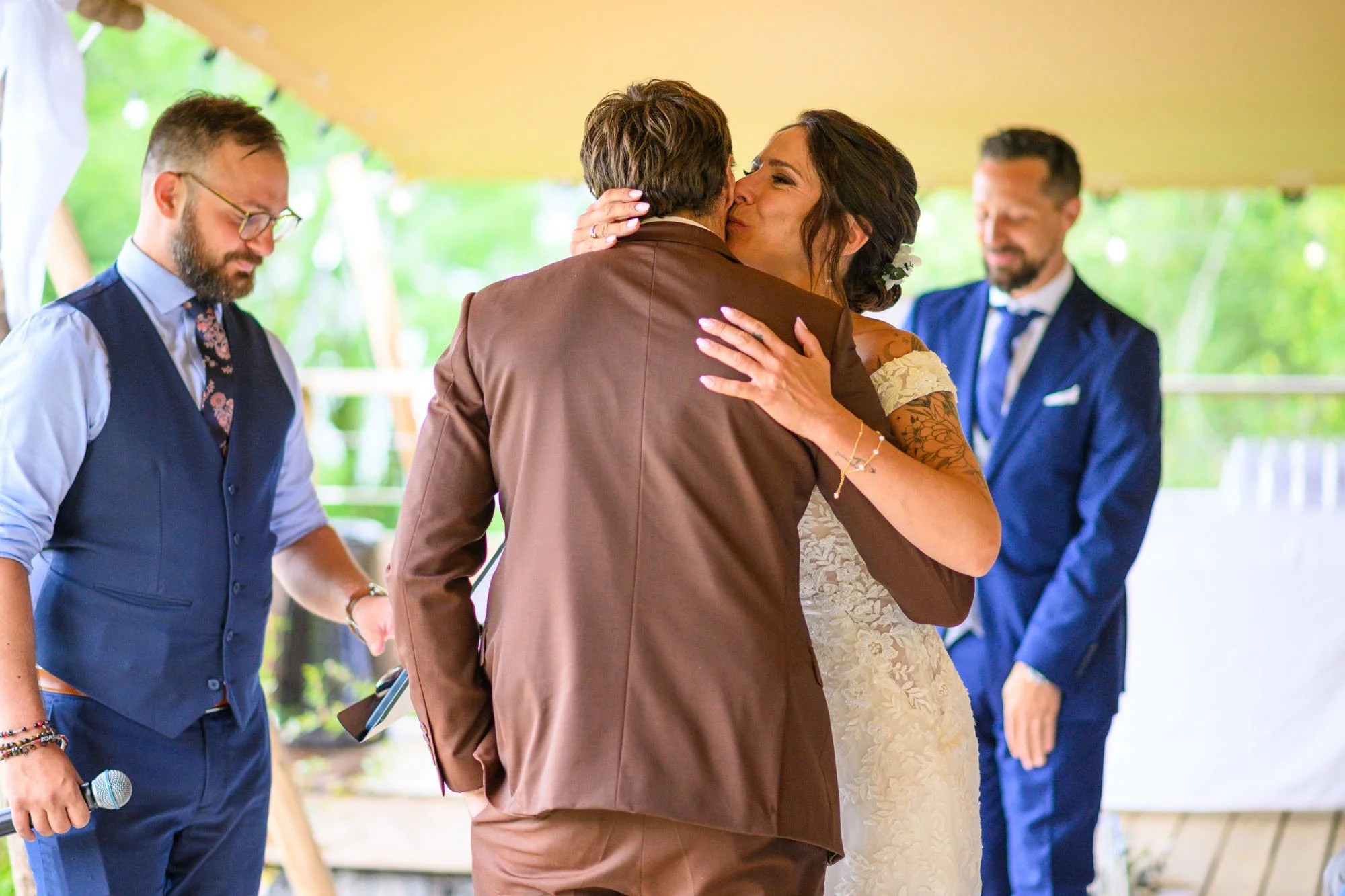 Mariage avec la mariée embrassant un homme en costume marron, entourée de deux hommes en costumes bleus, dans un cadre extérieur.