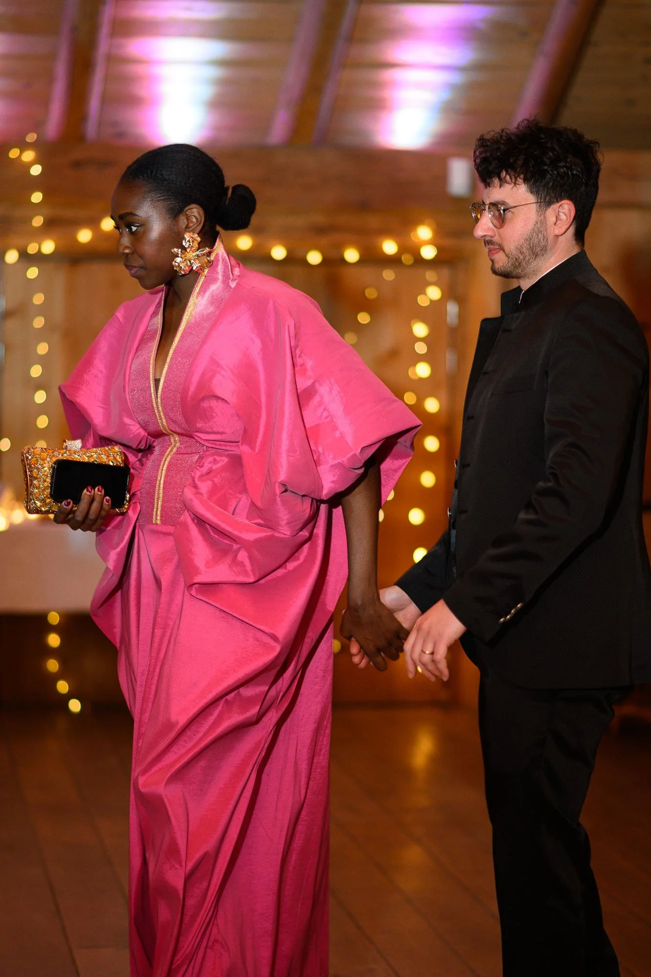 Deux personnes, une femme noire en robe rose et un homme blanc en costume noir, se tiennent par la main lors d'un événement, avec un fond de guirlandes lumineuses.