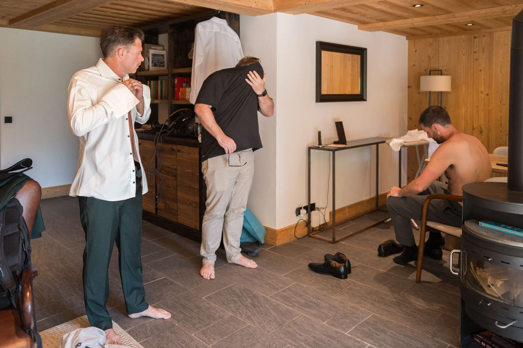 Une scène dans une pièce de style rustique avec quatre hommes. Deux d'entre eux sont debout, l'un en costume avec une chemise blanche, l'autre en t-shirt noir et pantalon beige. Le troisième homme, assis, est torse nu et regarde en bas. Il semble s'a