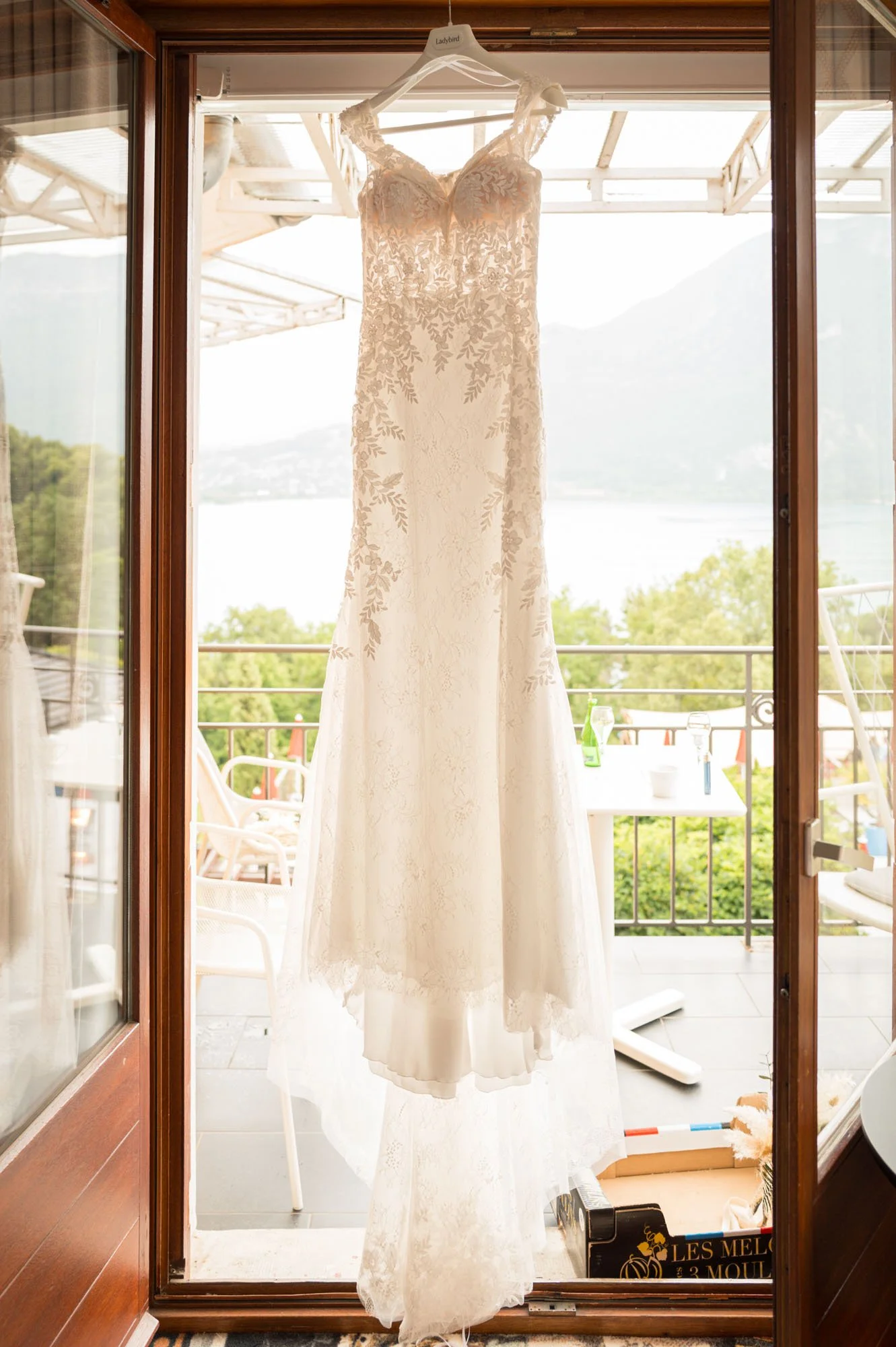Robe de mariage blanche en dentelle suspendue à une corde sur une véranda, avec vue sur un paysage montagneux et un lac en arrière-plan.