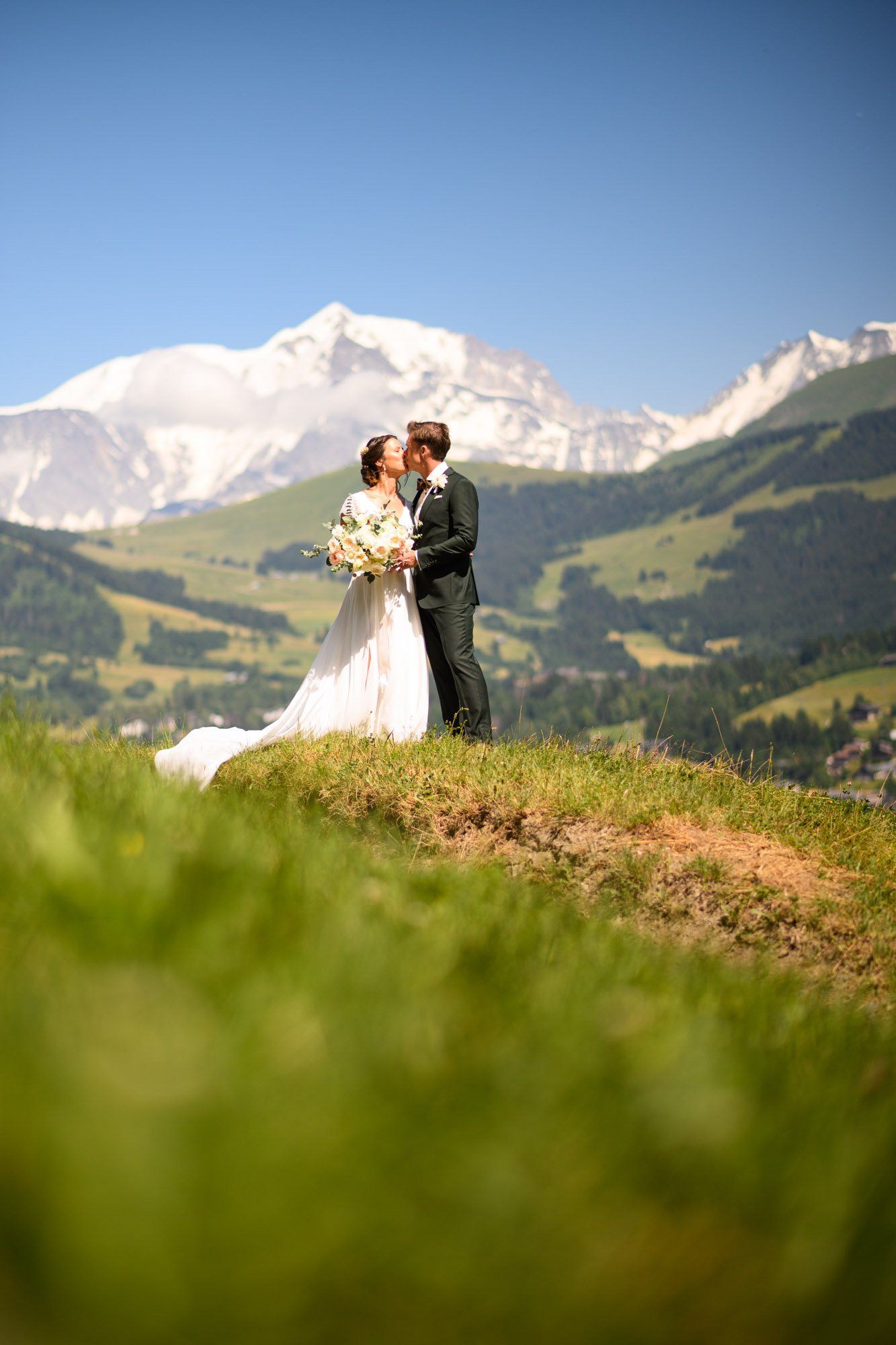 Un couple de mariés, un homme en costume noir et une femme en robe de mariée blanche, s'embrassant en face d'un paysage montagneux avec des sommets enneigés et un ciel bleu.