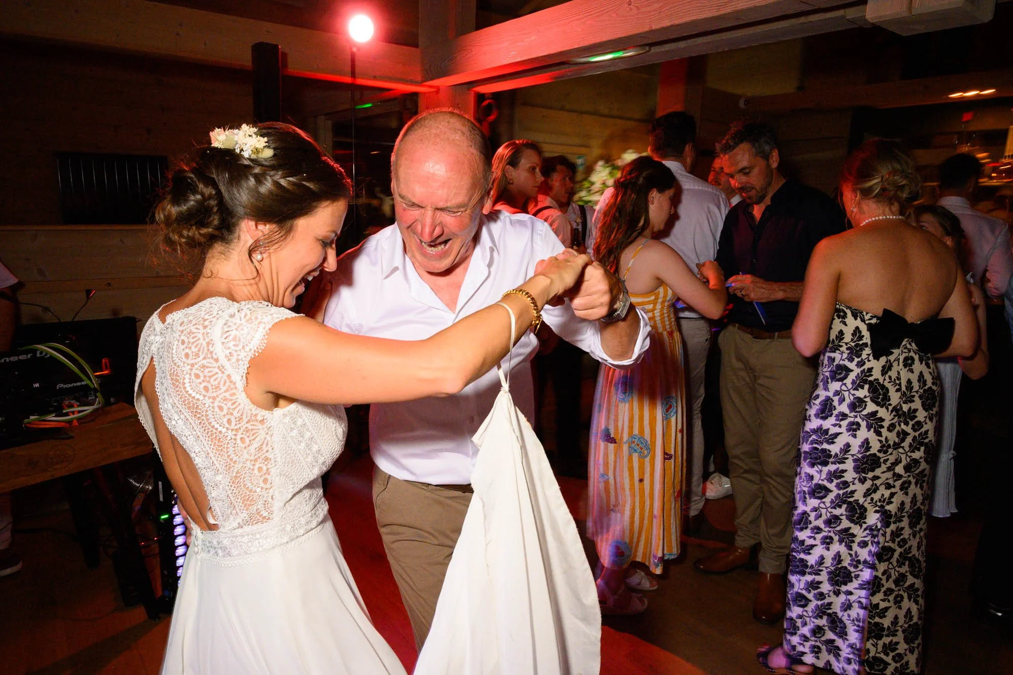 Gros plan sur une femme en robe de mariée et un homme âgé qui dansent ensemble, souriant, dans une ambiance de fête avec plusieurs autres personnes en arrière-plan.