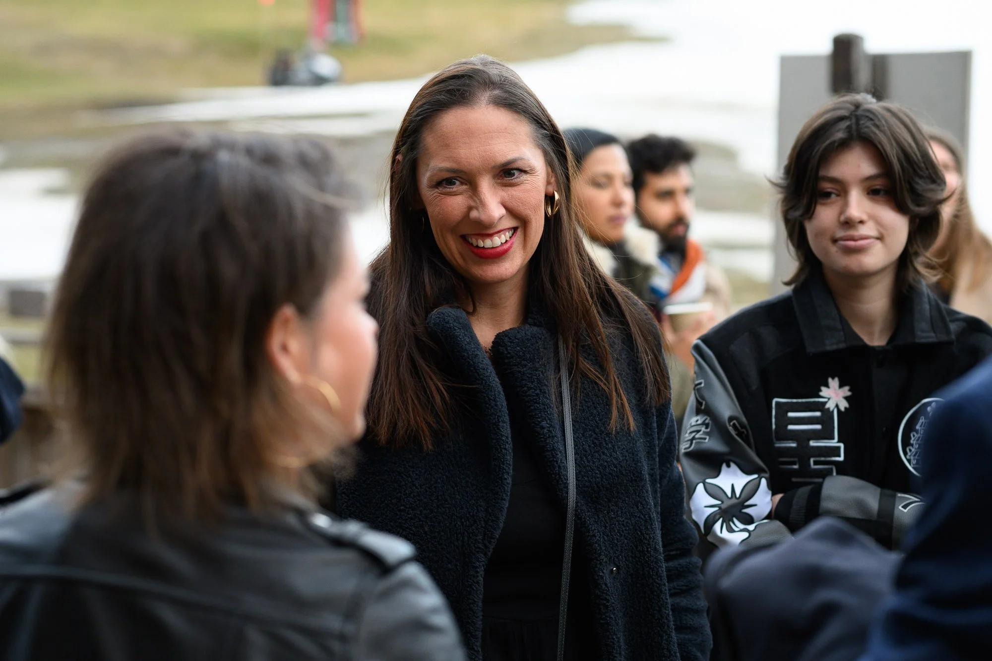 Groupe de personnes discutant à l'extérieur près d'un plan d'eau, avec une femme souriante au centre en noir, entourée de jeunes et d'autres adultes.