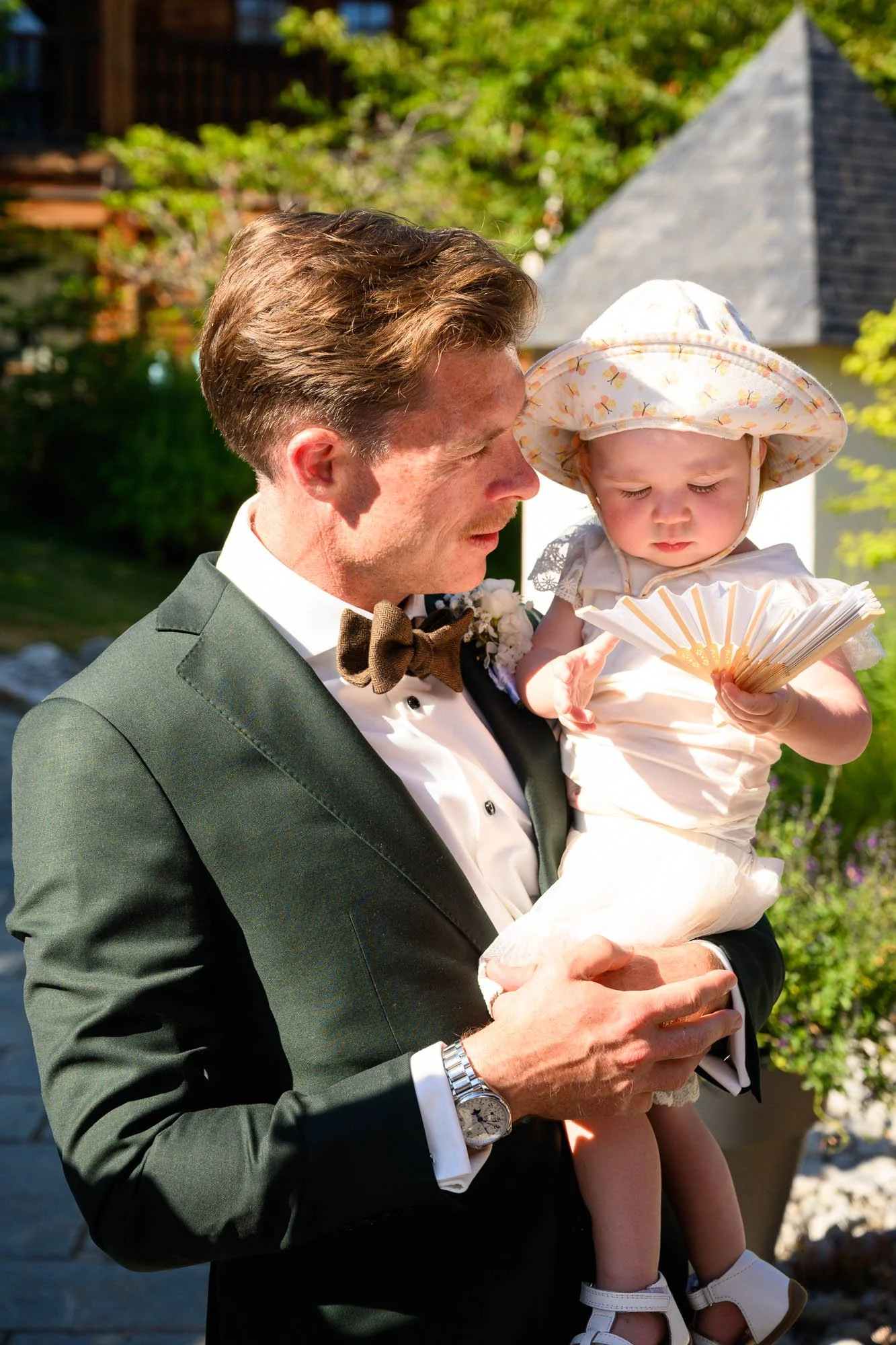 Un homme en costume noir, portant une veste élégante et une cravate en nœud papillon, tient une petite fille habillée en blanc avec un chapeau de soleil, lors d'une cérémonie en plein air.