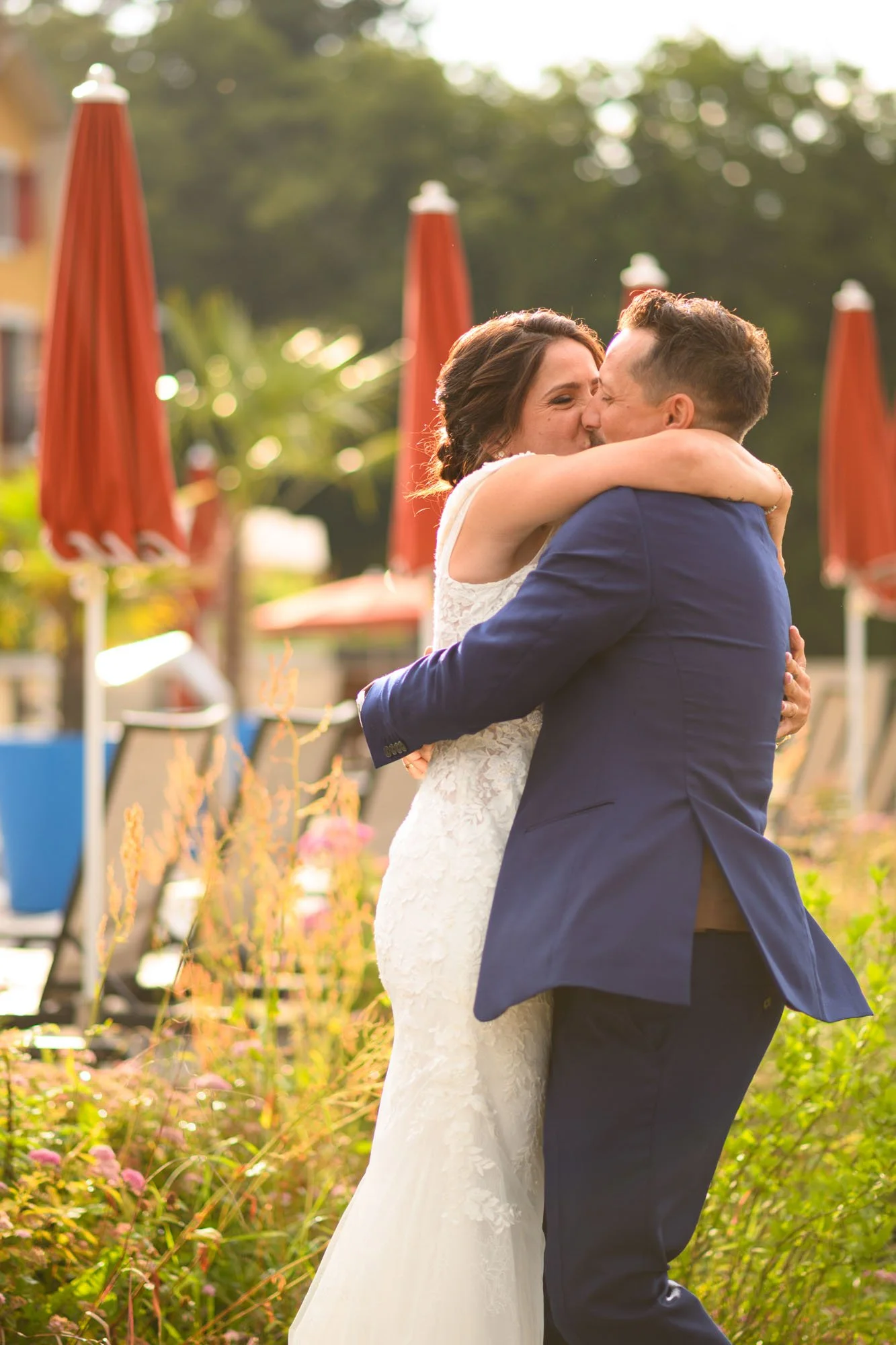Un couple en costume de mariage s'embrasse en plein air, entouré de parasols rouges et de végétation.