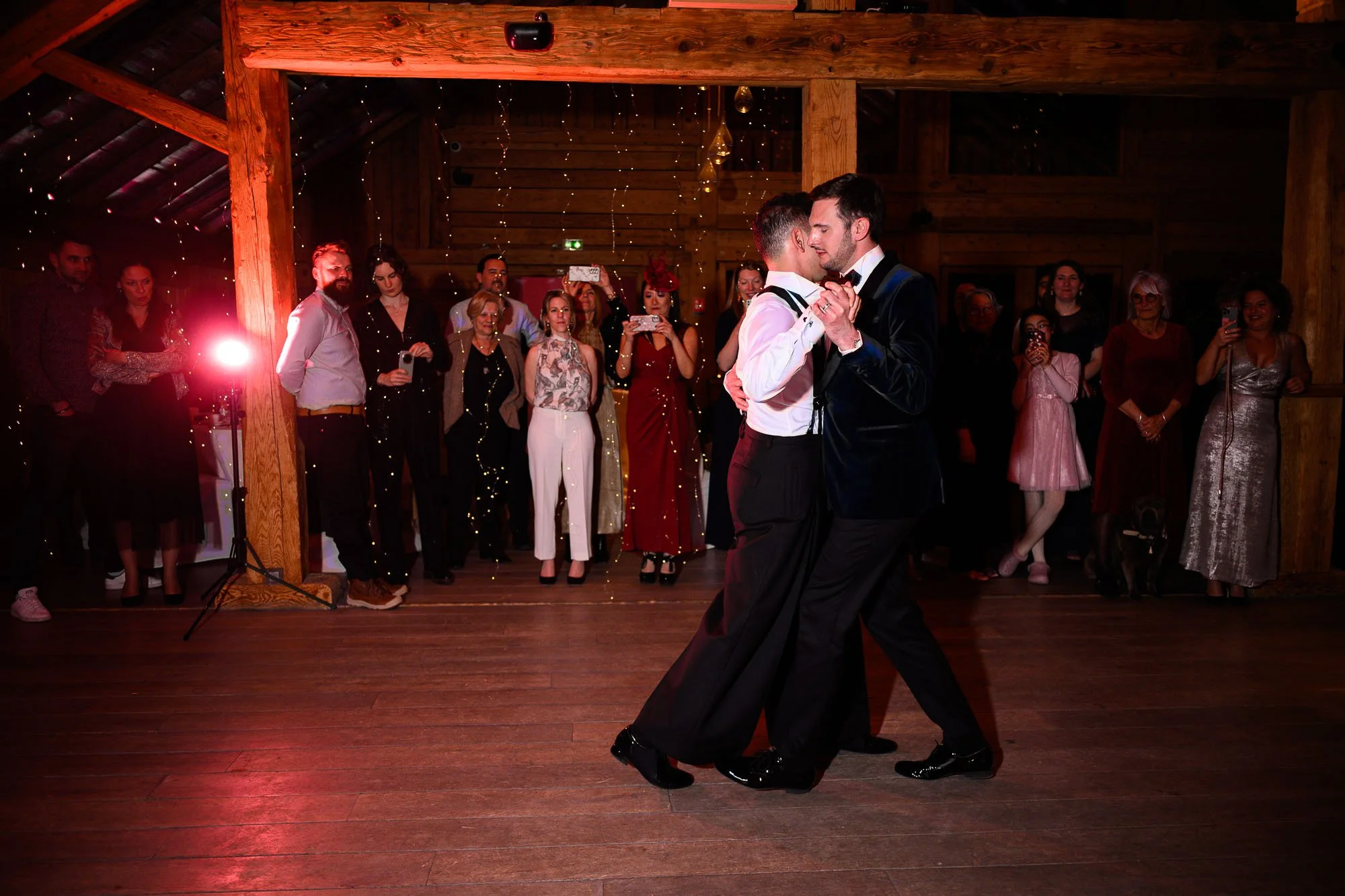 Deux hommes dans un costume de mariage dansent en ferme proximité lors d'une réception dans une salle en bois, entourés de plusieurs invités qui regardent et prennent des photos.