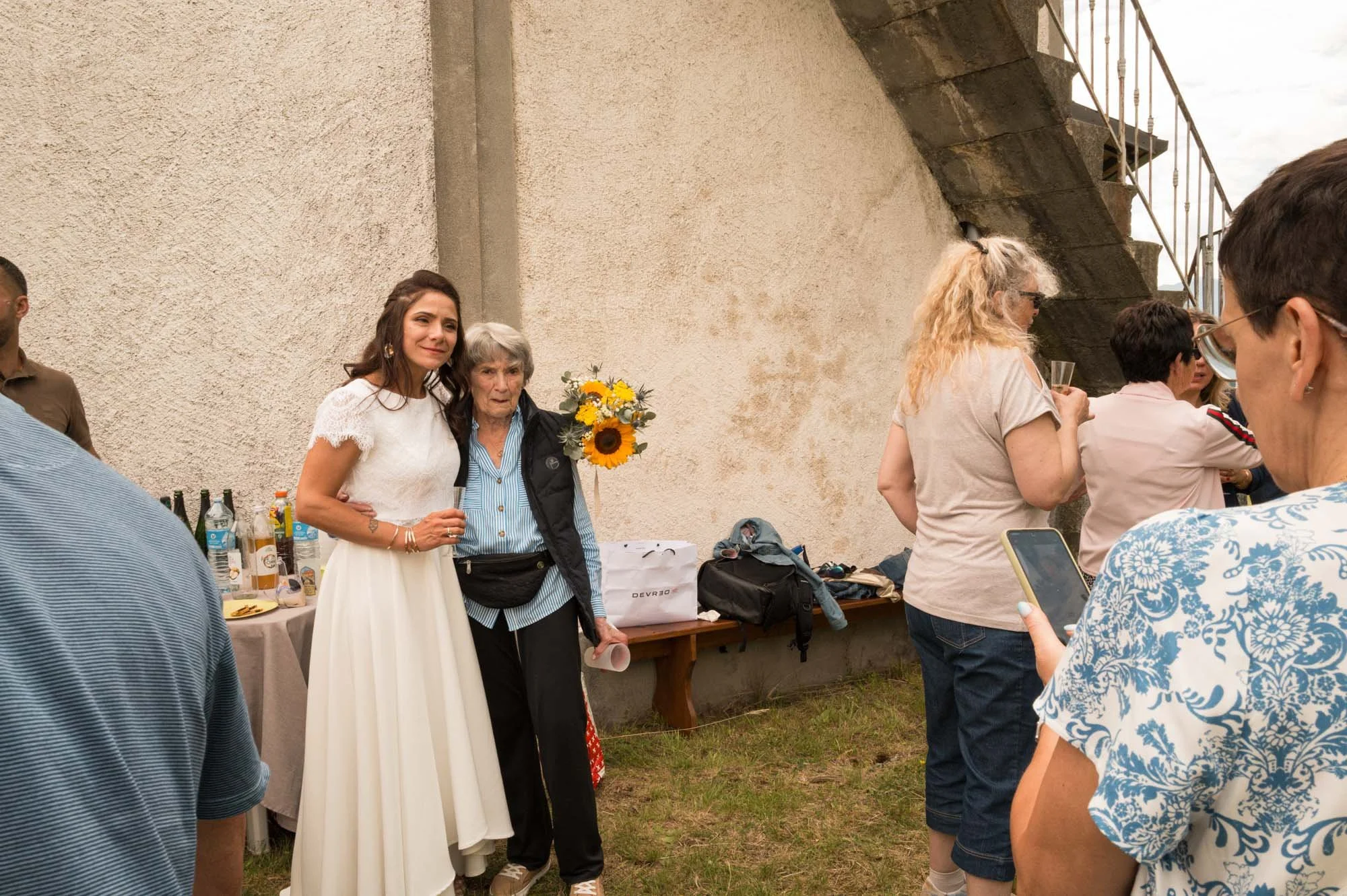 Une jeune femme en robe blanche posant avec une femme âgée tenant un bouquet de tournesols lors d'une fête ou d'un rassemblement en plein air.