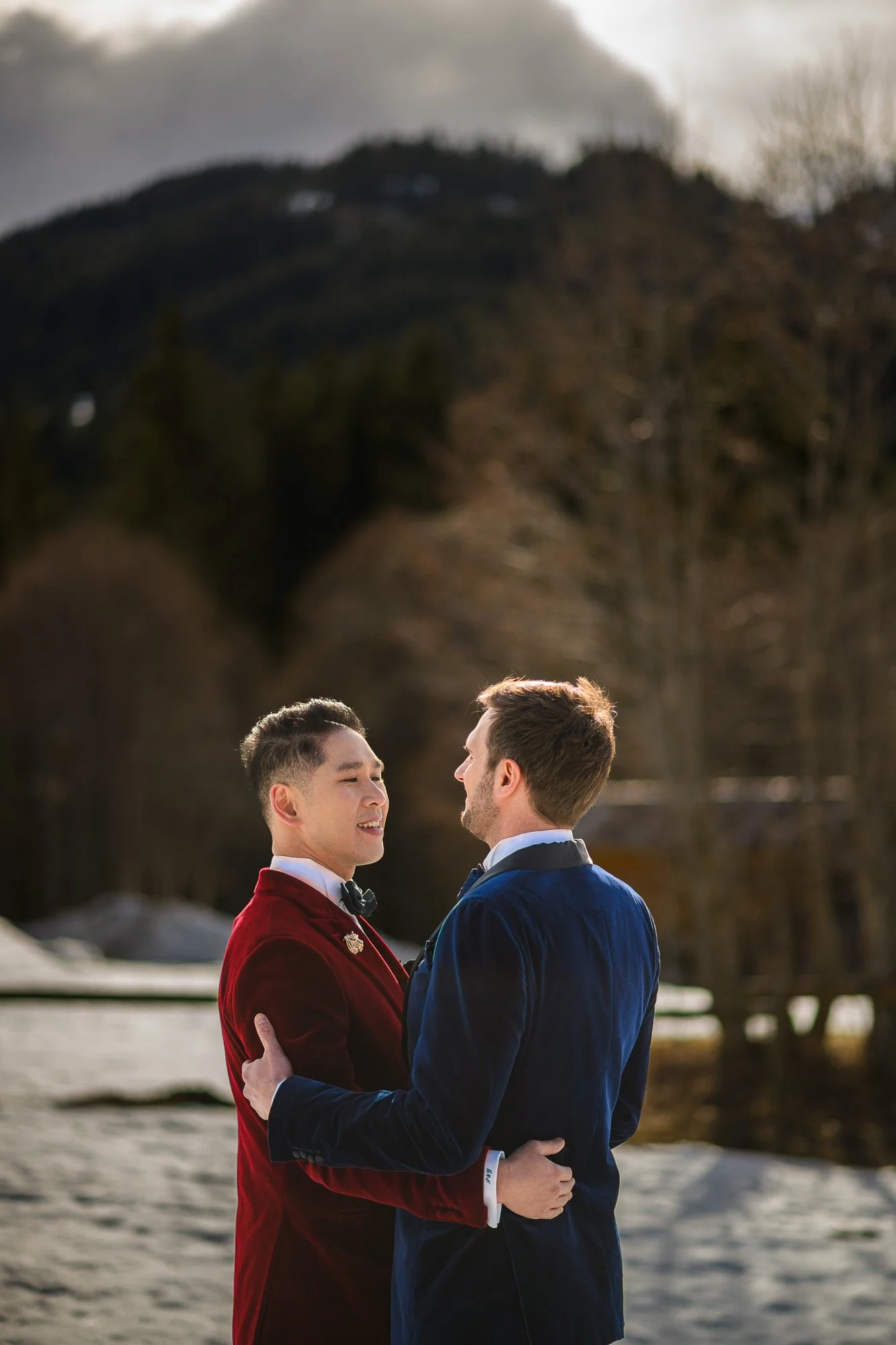 Deux hommes habillés en costumes de velours, une en rouge et l'autre en bleu, s'embrassent devant un paysage de montagnes et d'arbres en automne, en plein air.