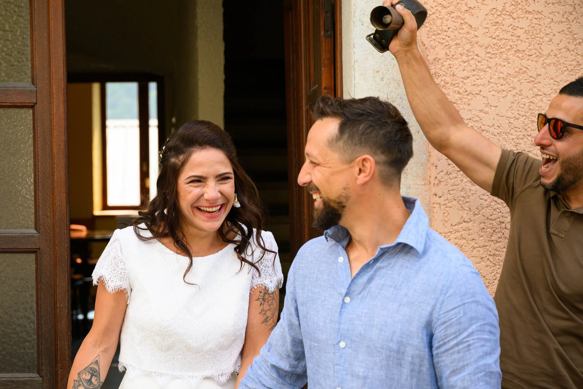 Trois personnes souriantes et riant, une femme avec des tatouages sur le bras, un homme en chemise bleue, un autre homme avec des lunettes de soleil, dans une scène joyeuse en extérieur.
