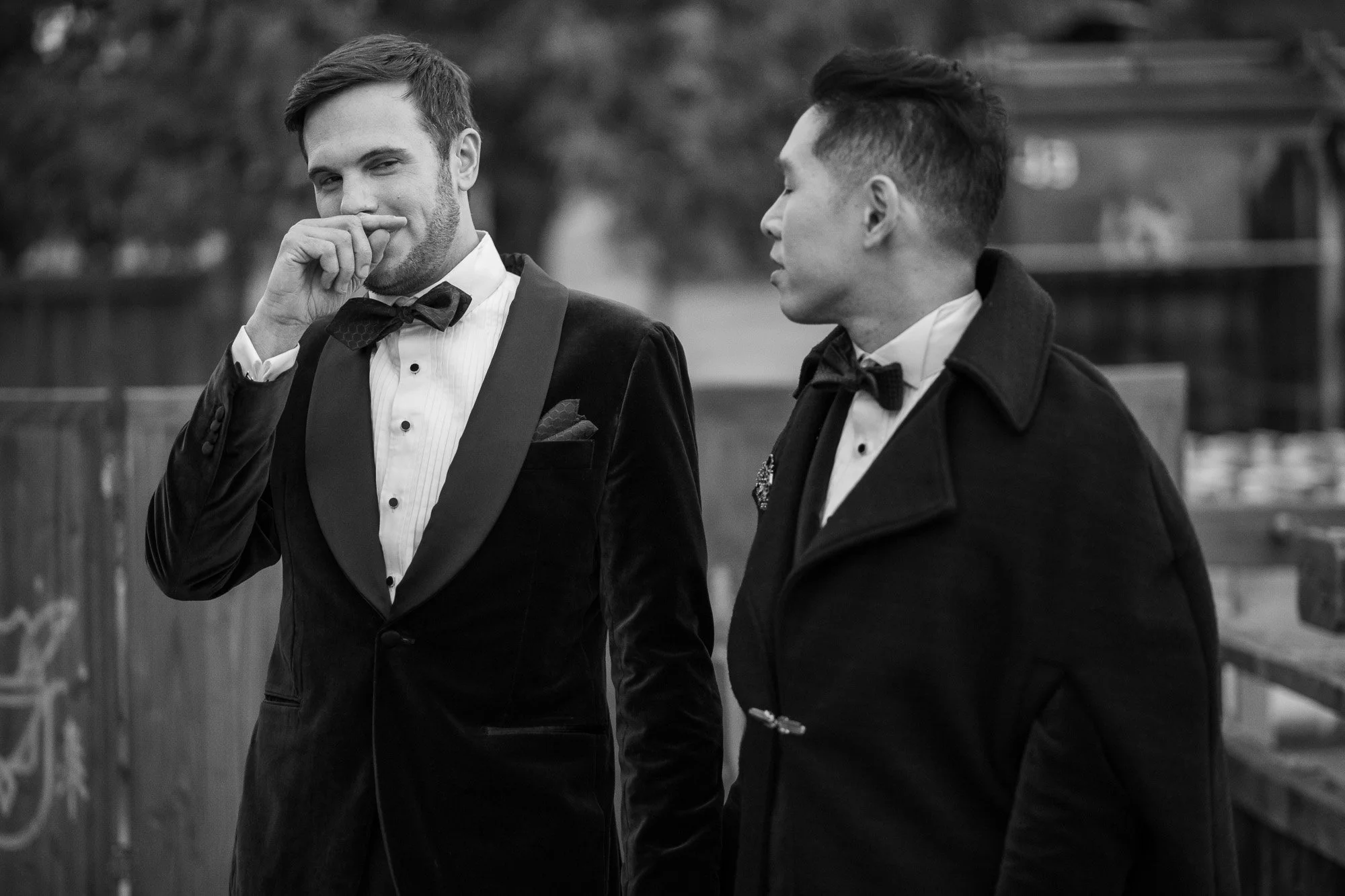 Deux hommes en costumes sombres, l'un avec un nœud papillon, discutent dehors, en noir et blanc.