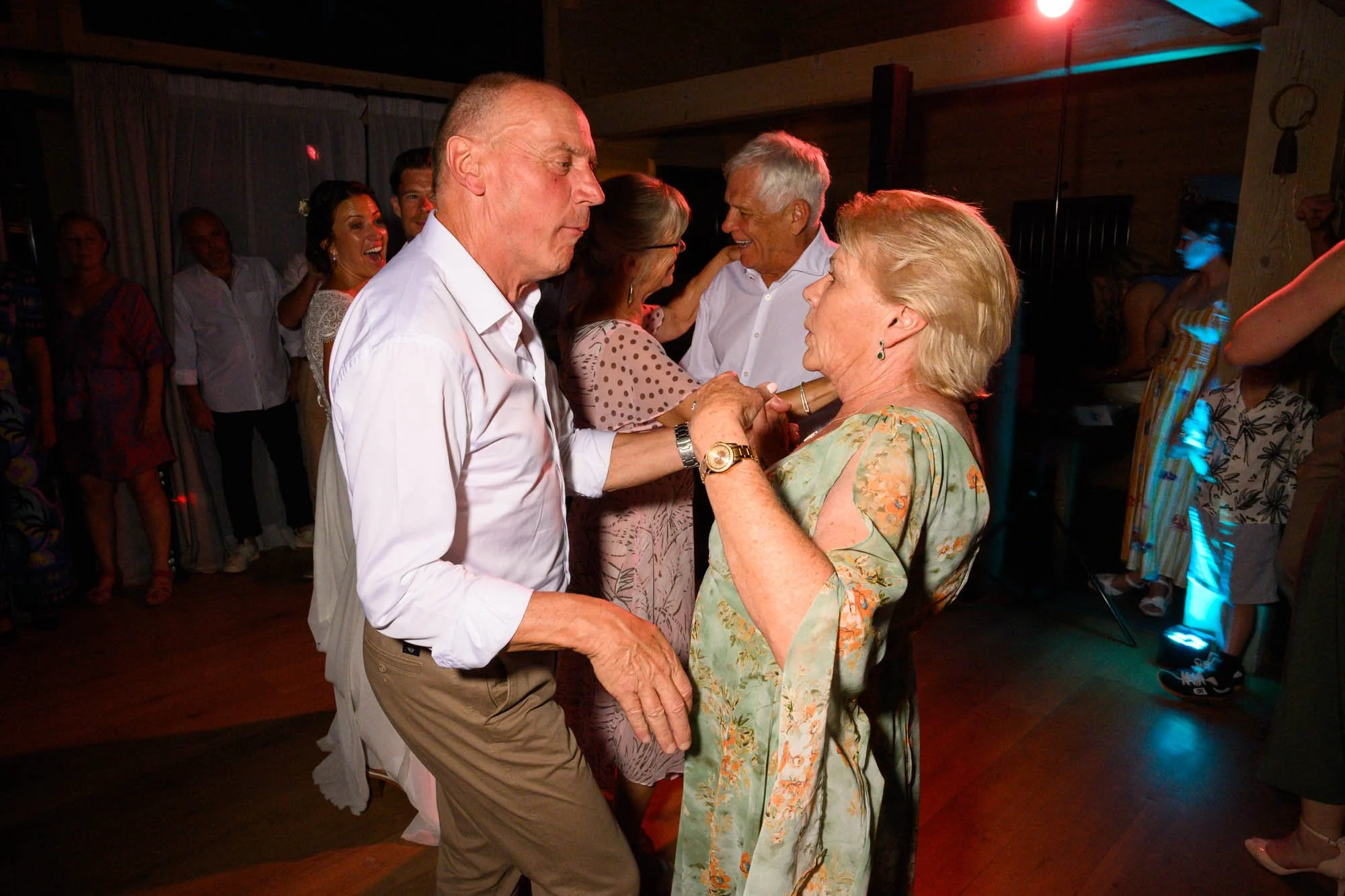 Des personnes âgées dansent lors d'une fête, illuminée par des lumières colorées, dans une pièce avec un parquet en bois.