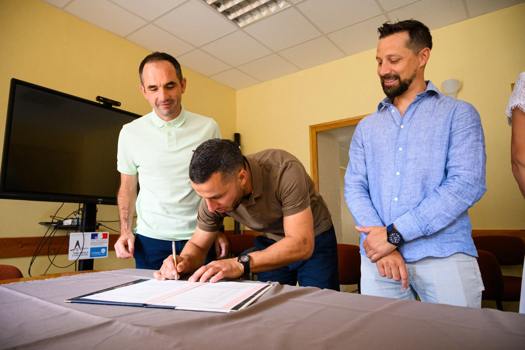 Un homme est en train de signer un document sur une table, entouré de trois autres hommes qui regardent. L'atmosphère semble formelle et professionnelle.