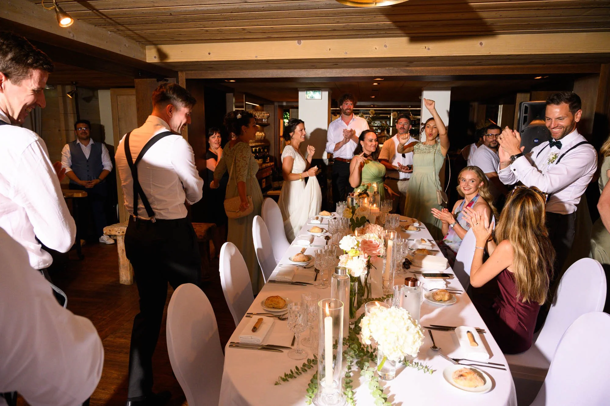 Un groupe de personnes lors d'un repas de mariage, certaines applaudissant et d'autres debout autour d'une table décorée avec des fleurs et des bougies.