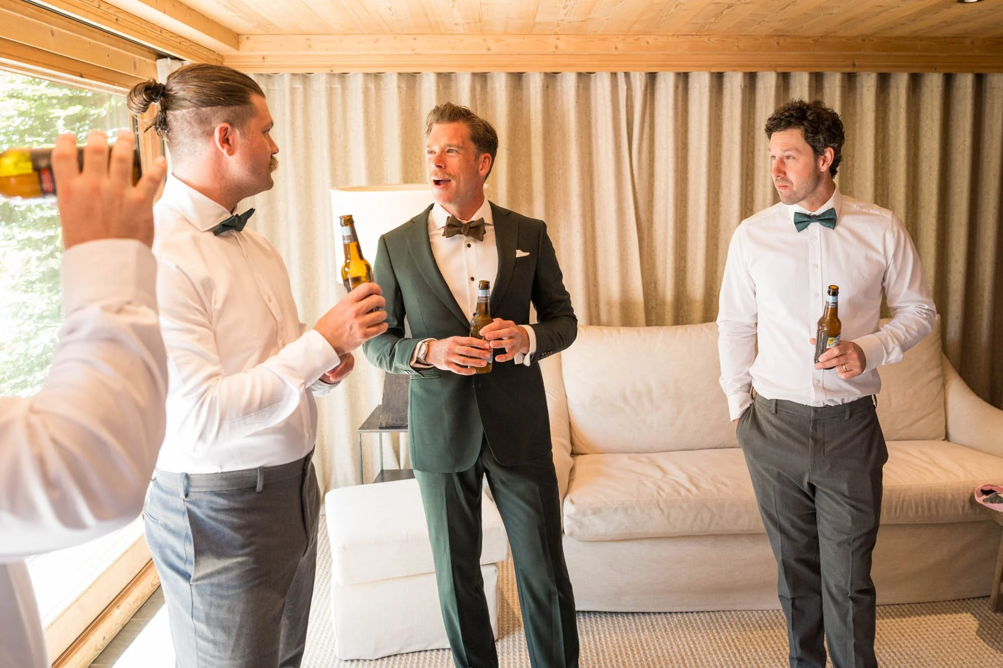 Quatre hommes en costumes habillés, discutant et tenant des bières dans un salon avec des rideaux et un canapé beige.