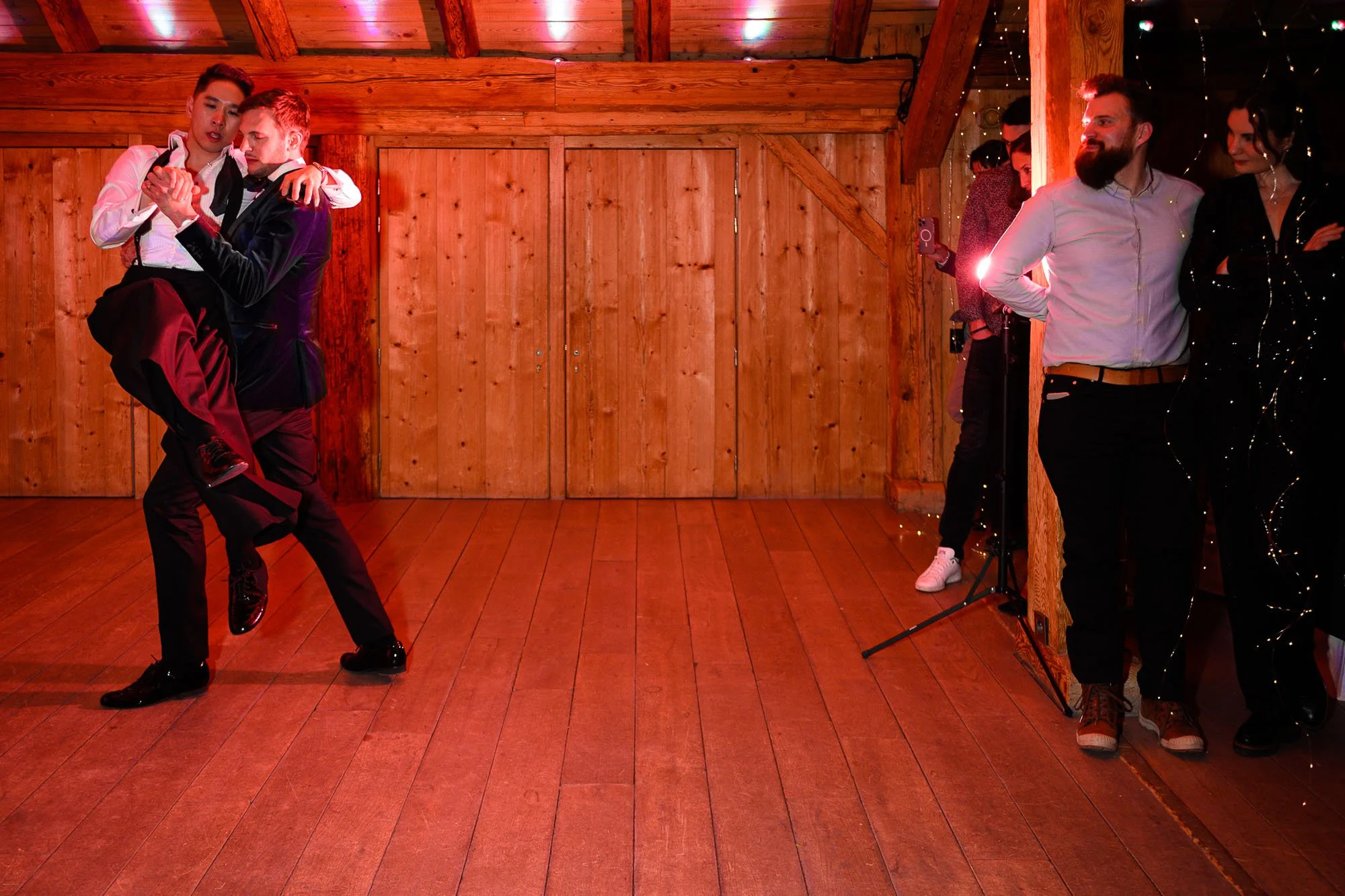 Deux jeunes hommes dansants lors d'une soirée dans une salle en bois, avec un groupe de spectateurs observant à droite, dont un homme avec une barbe et une femme en noir