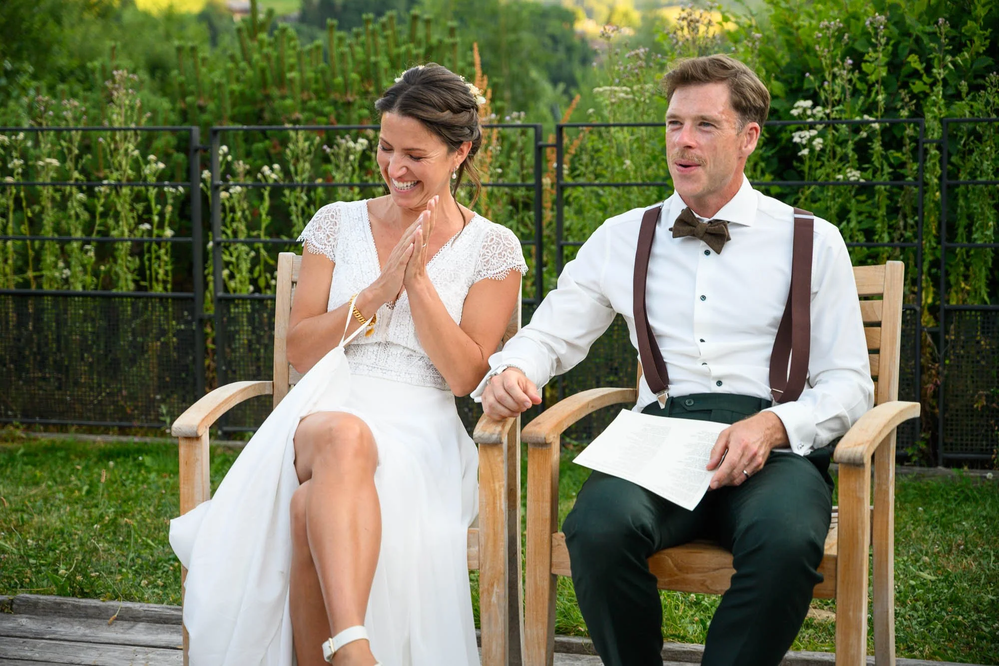 Un couple lors d'une cérémonie de mariage assis sur des chaises en bois à l'extérieur, avec un fond de végétation verte. La femme porte une robe blanche et sourit, tandis que l'homme, en chemise blanche, porte un nœud papillon et des bretelles, et ti