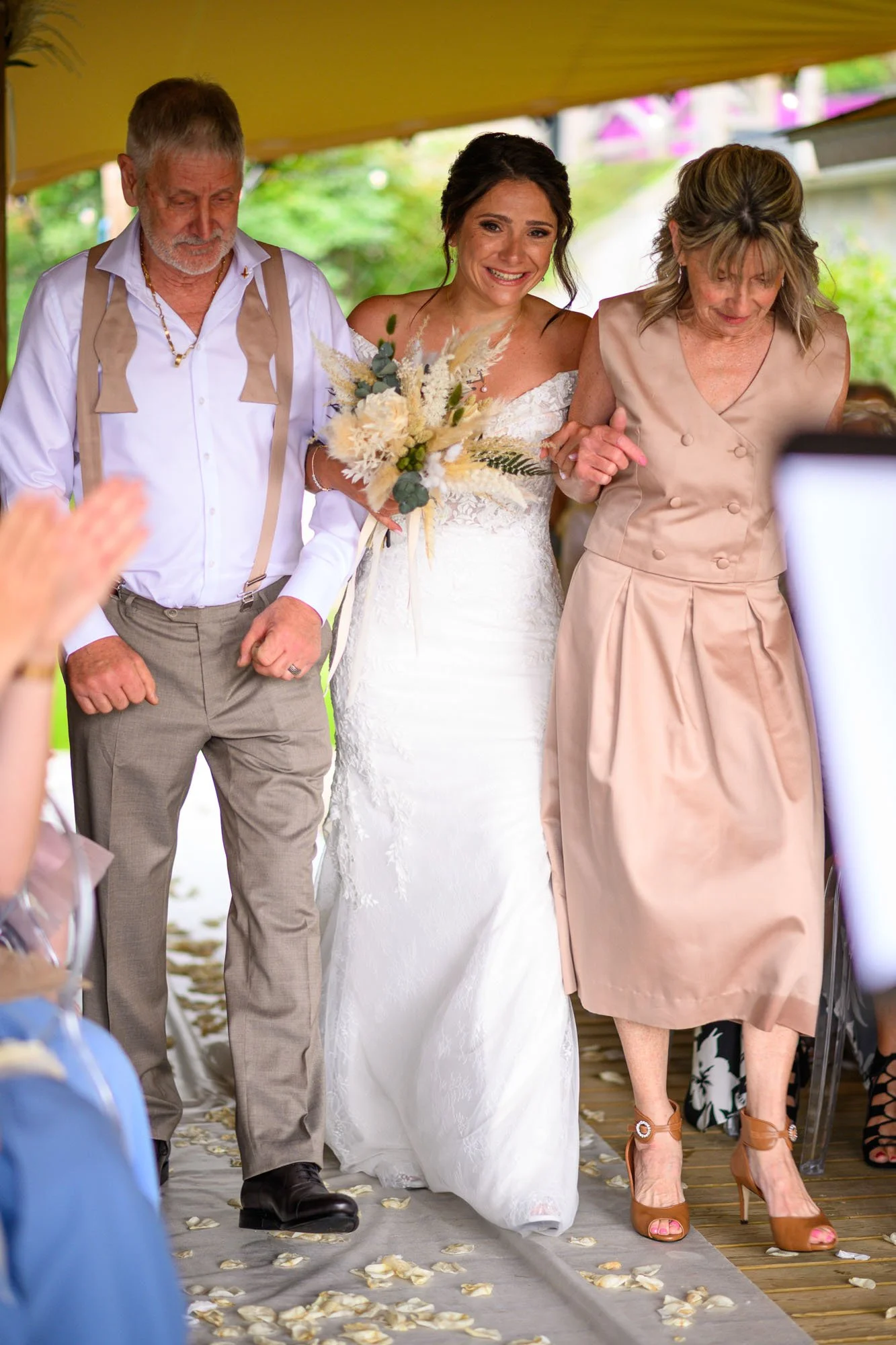 Une mariée en robe blanche est aidée à marcher par ses parents lors d'une cérémonie de mariage, avec des invités en arrière-plan.