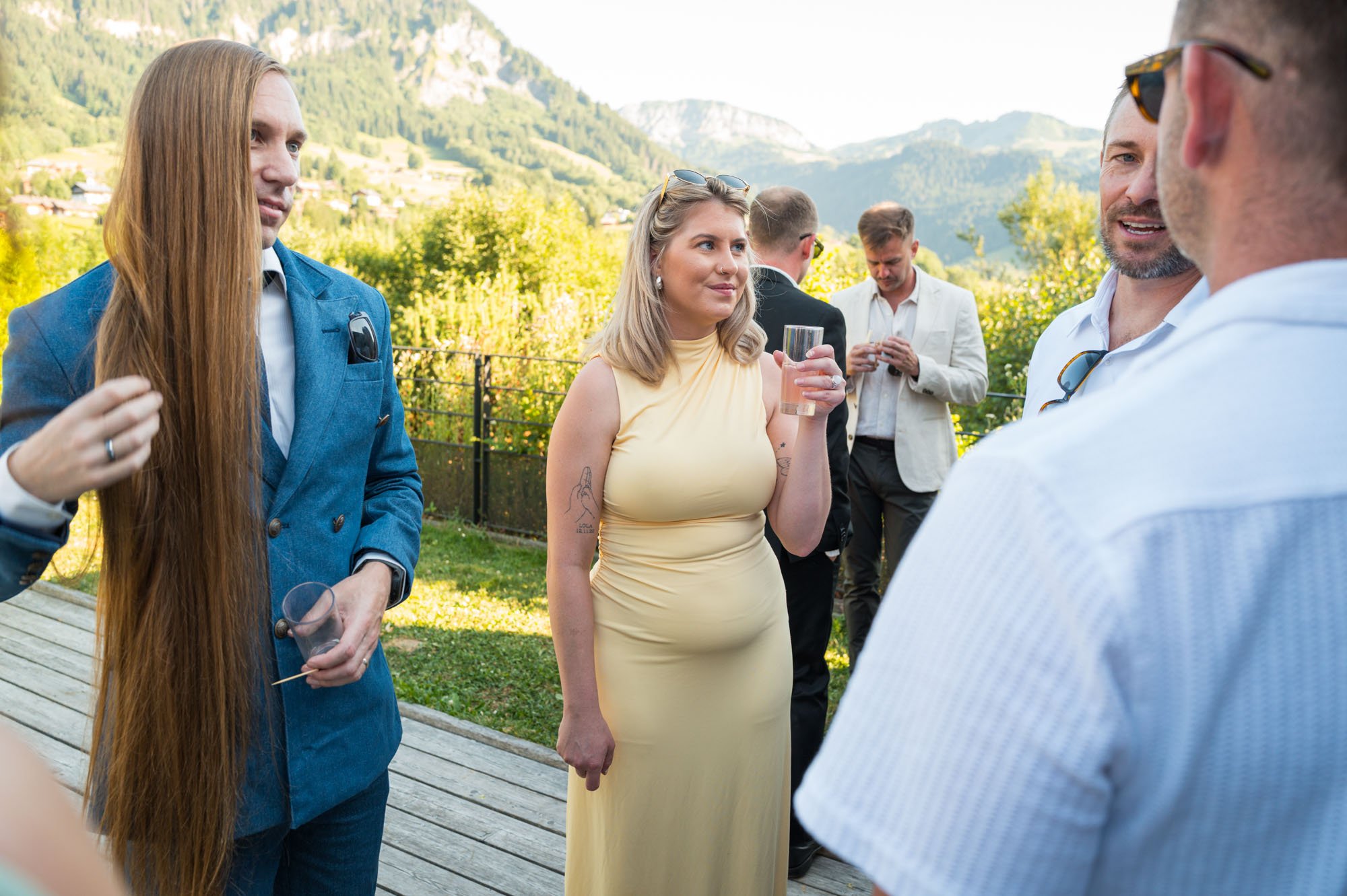 Groupe de personnes lors d'une réception en extérieur avec vue sur la montagne, échangeant des conversations et tenant des verres.