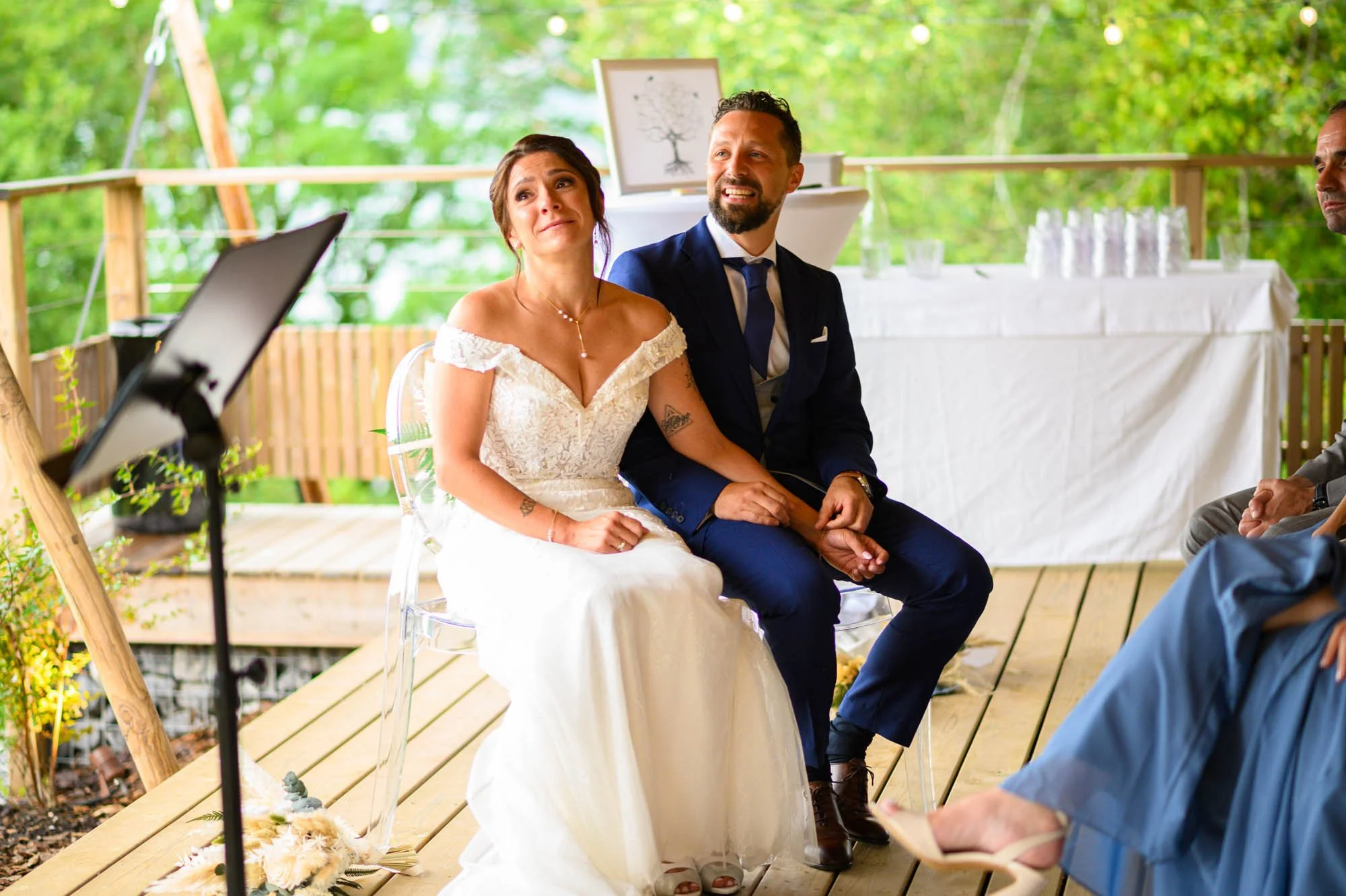 Un couple de mariés assis lors de leur cérémonie de mariage en plein air, entourés de verdure.
