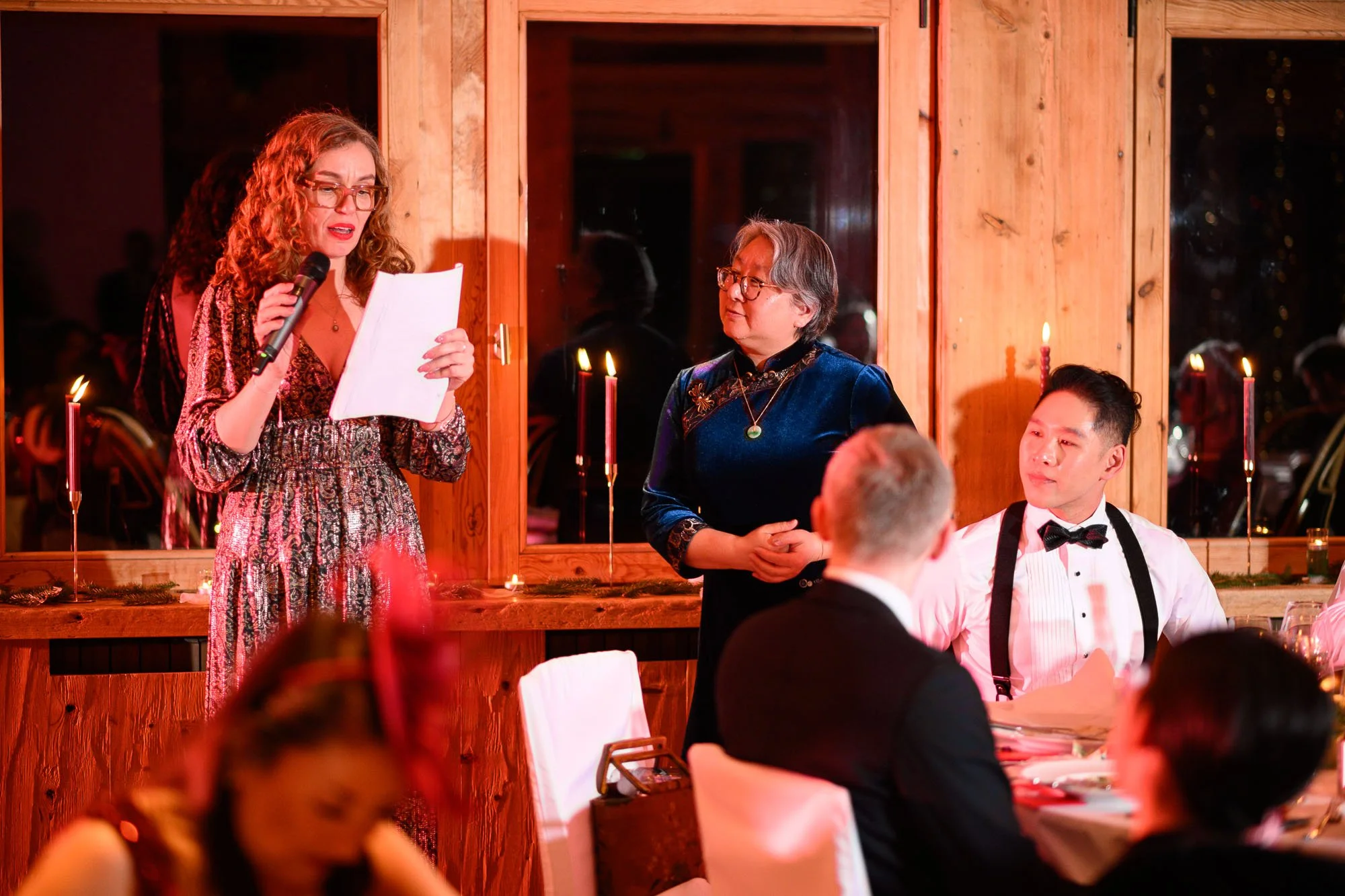 Une femme avec des cheveux roux portant une robe à motifs lit un discours lors d'un dîner de gala, devant une autre femme en velours bleu et un jeune homme en smoking blanc avec nœud papillon, dans une salle décorée de bougies