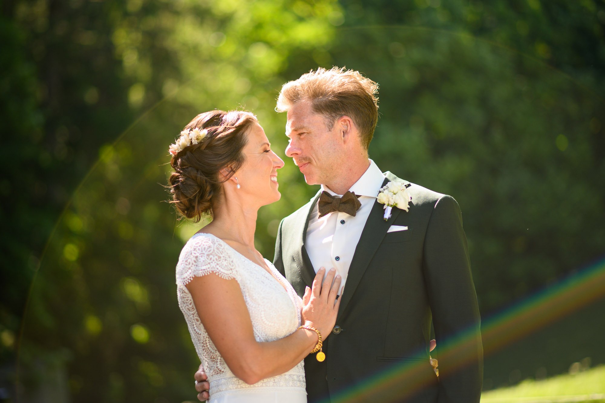 Un couple en mariage, la femme en robe blanche avec un bouquet blanc, l'homme en costume noir avec papillon, se regardant dans un cadre naturel en plein air.
