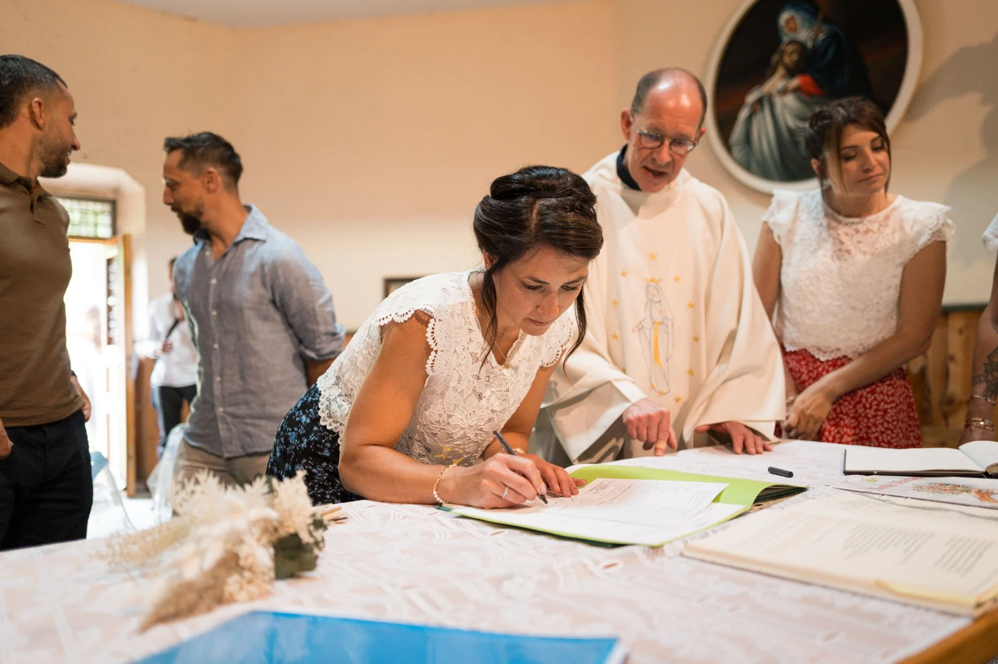 Une femme signe un registre lors d'une cérémonie religieuse, entourée d'un prêtre et d'autres personnes, dans une salle décorée.