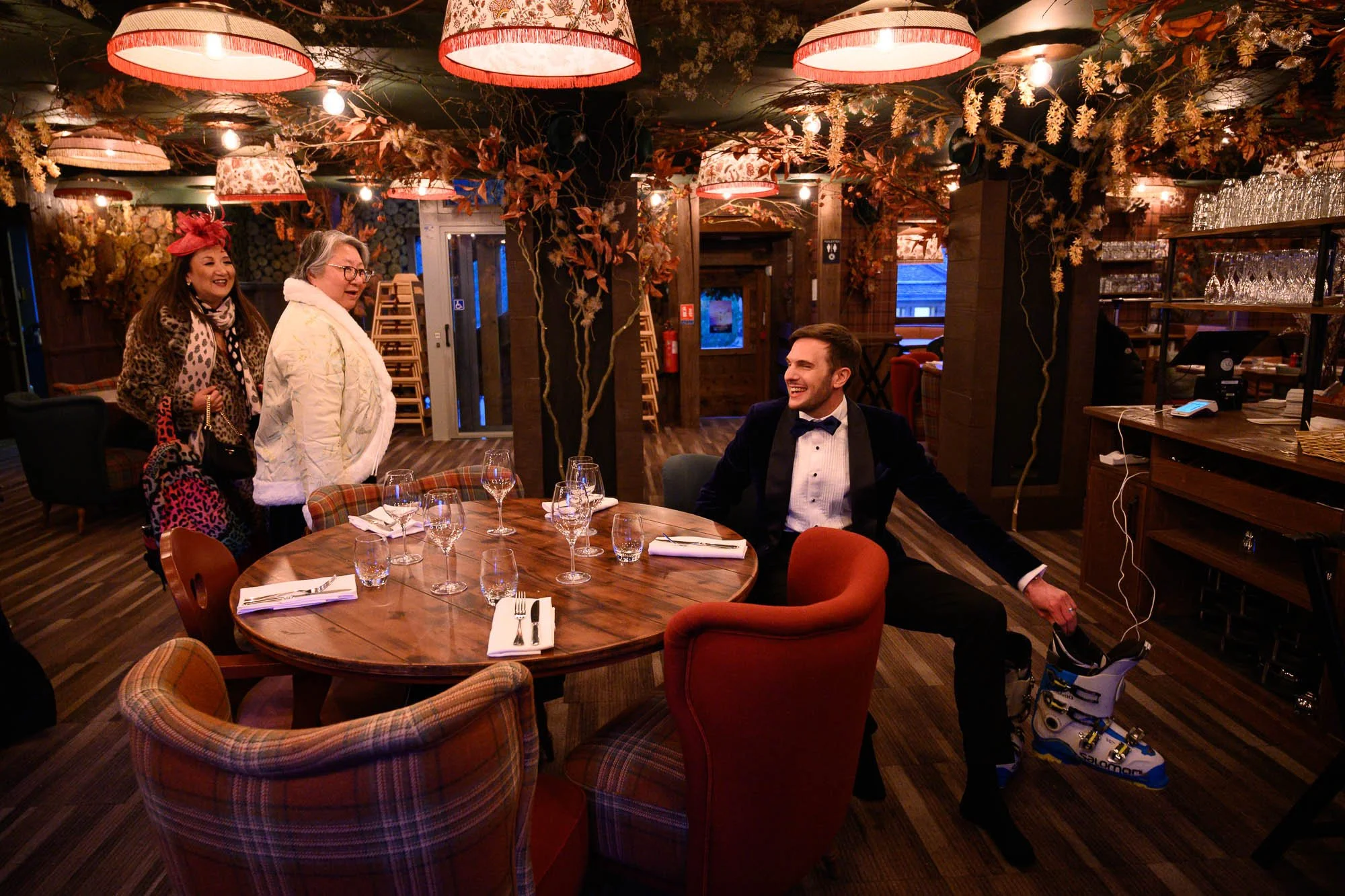 Une scène dans un restaurant avec un homme en smoking assis à une table, deux femmes debout parlant avec lui, le décor est chaleureux avec des lumières tamisées, des plantes et des décorations automnales.