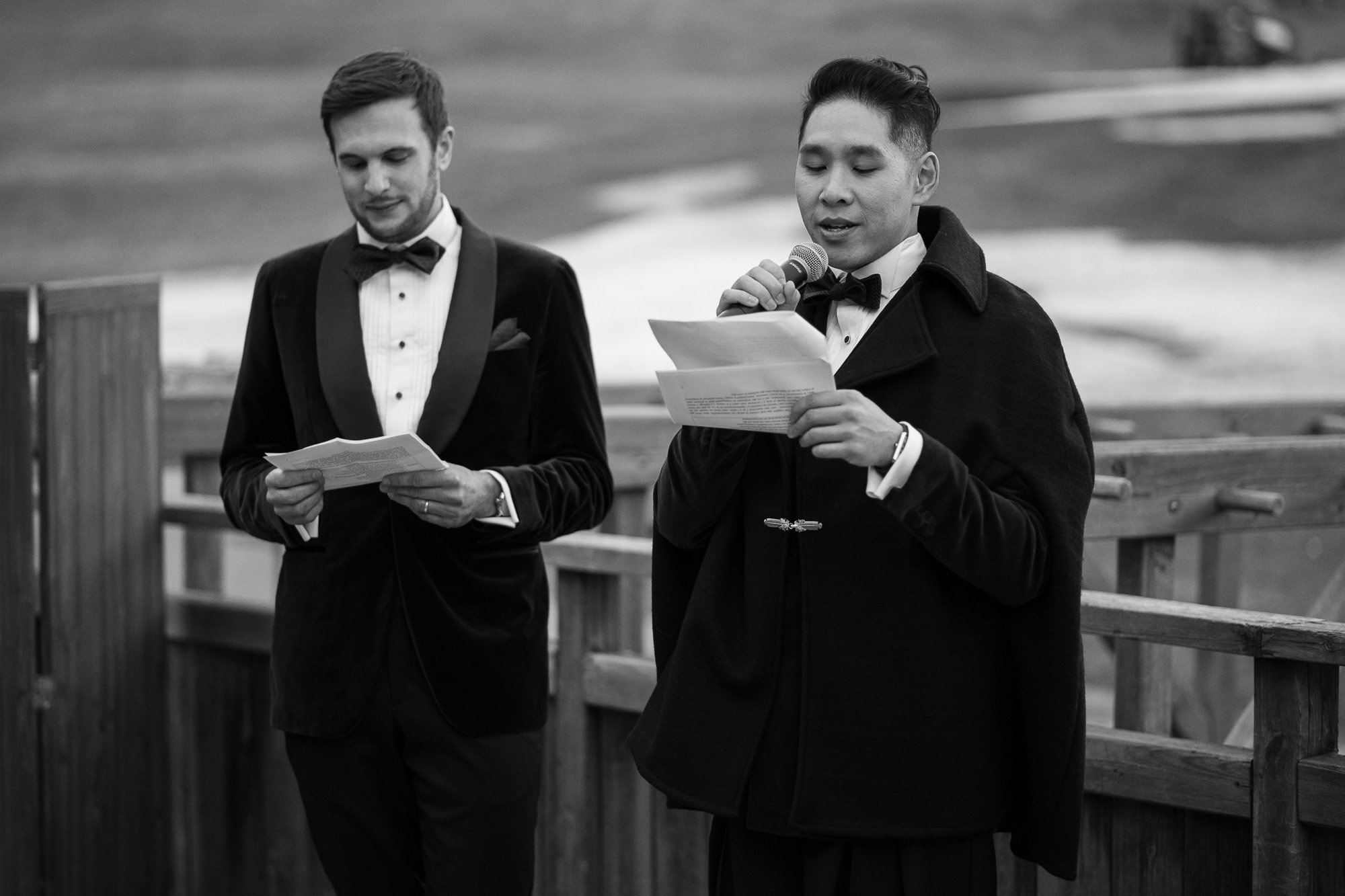 Deux hommes habillés en costume et smoking, en train de lire des textes lors d'une cérémonie en plein air, avec un fond de terrain et une clôture en bois.