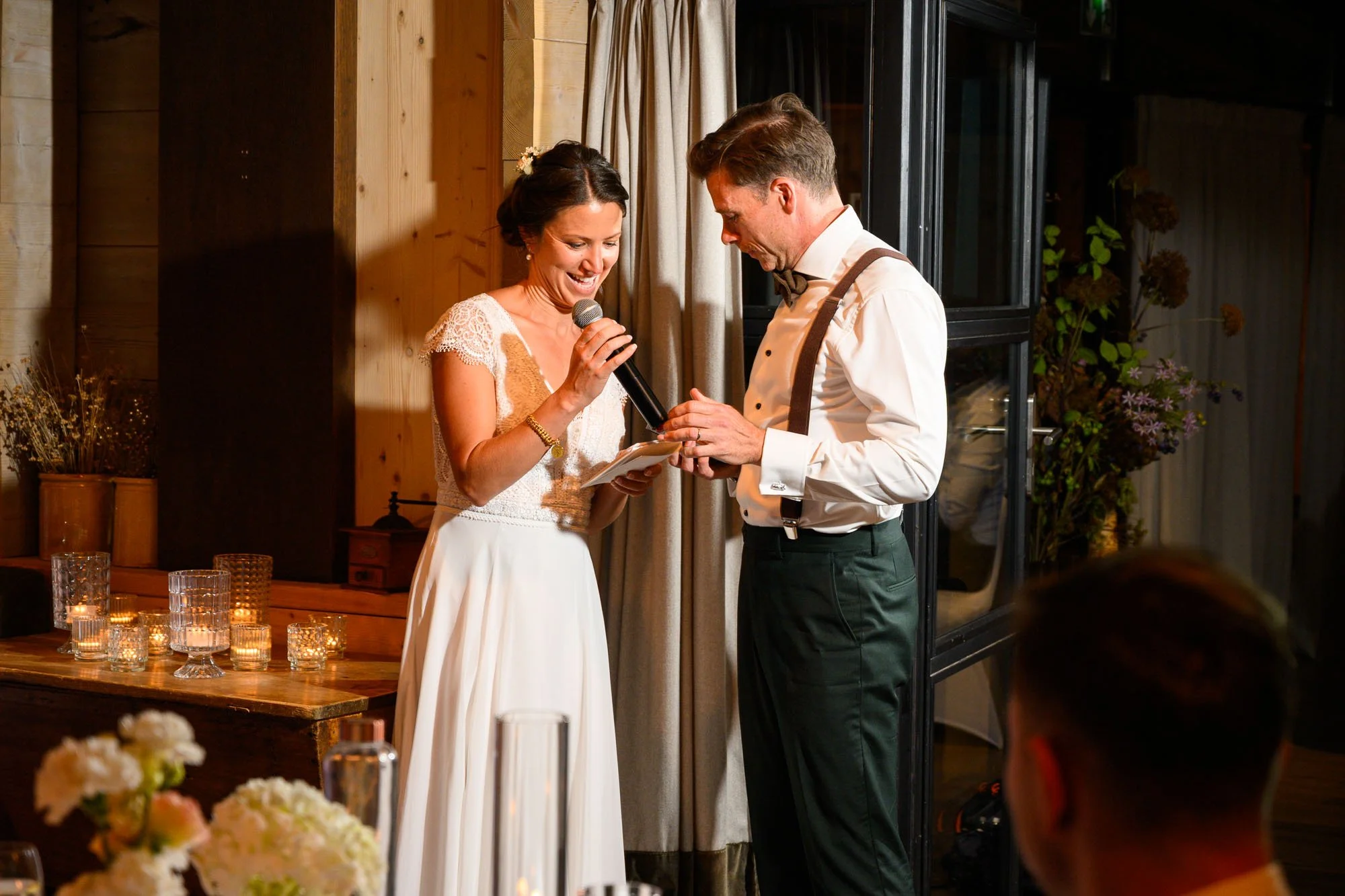 Un couple échange des bagues lors de leur mariage, la mariée tient un micro et le marié porte une chemise blanche avec un nœud papillon, dans une ambiance chaleureuse avec des bougies et des fleurs
