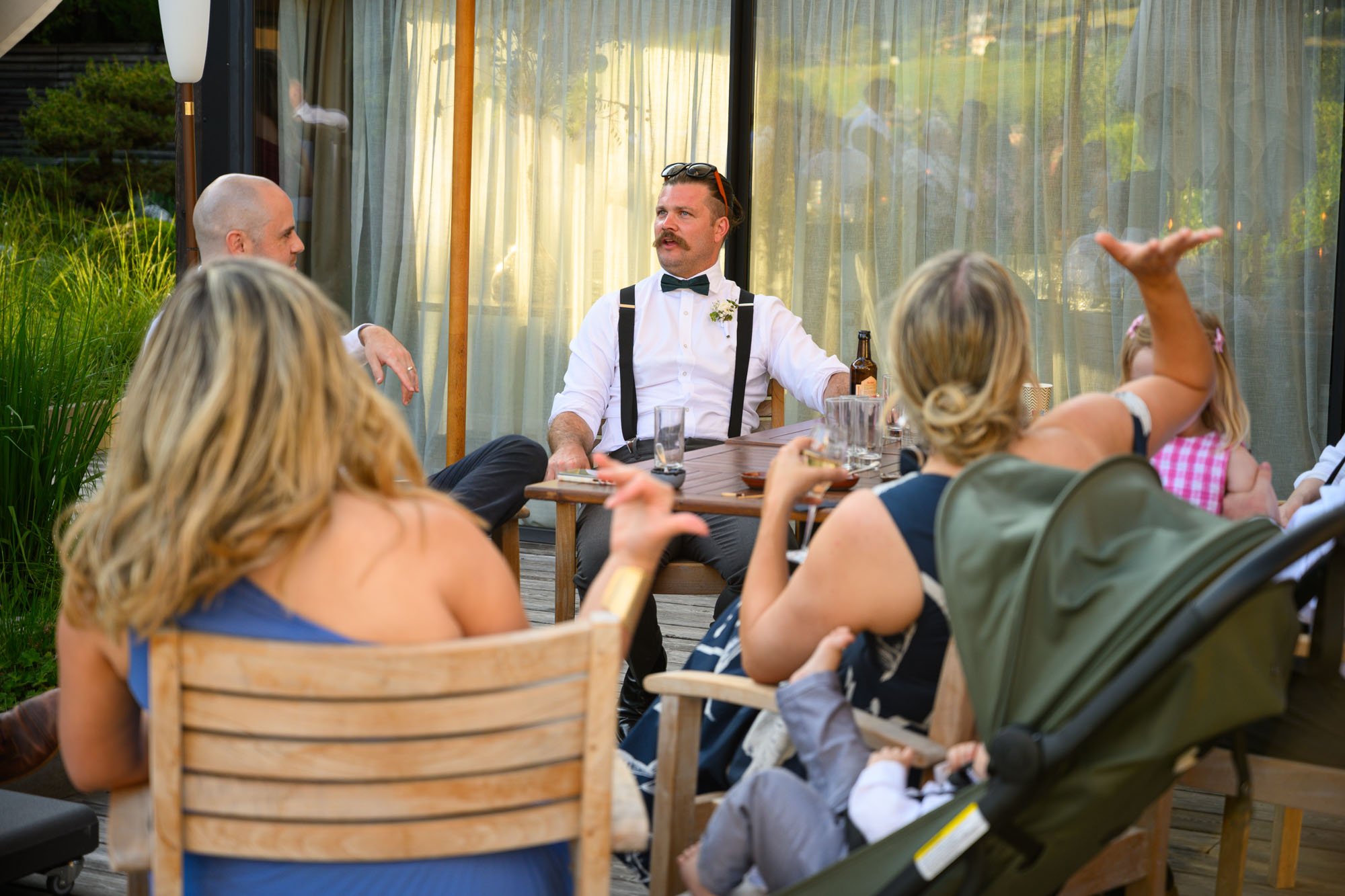 Groupe de personnes assises autour d'une table lors d'une fête en plein air, avec un homme en style vintage parlant, et des enfants, un bébé en poussette