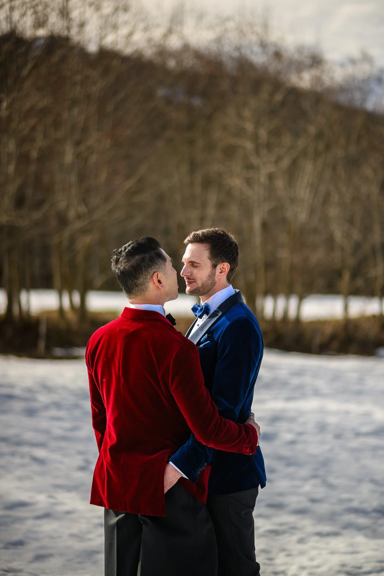 Deux hommes habillés en costumes de mariage, l'un en rouge et l'autre en bleu, s'embrassant tendre face à face dans un paysage hivernal avec des arbres nus en arrière-plan.