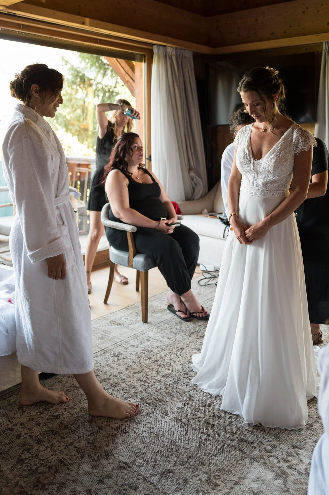 Une mariée en robe blanche se prépare dans une chambre avec plusieurs femmes, une en peignoir blanc, une autre assise sur une chaise, toutes entourant la mariée.