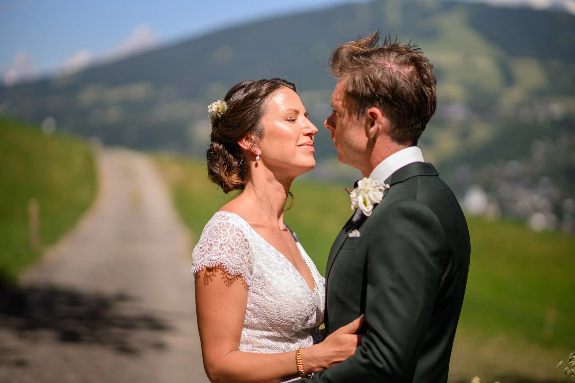 Un couple en robe de mariage et costume se tient face à face, sourire, en plein air avec un chemin longeant une colline verte en arrière-plan.