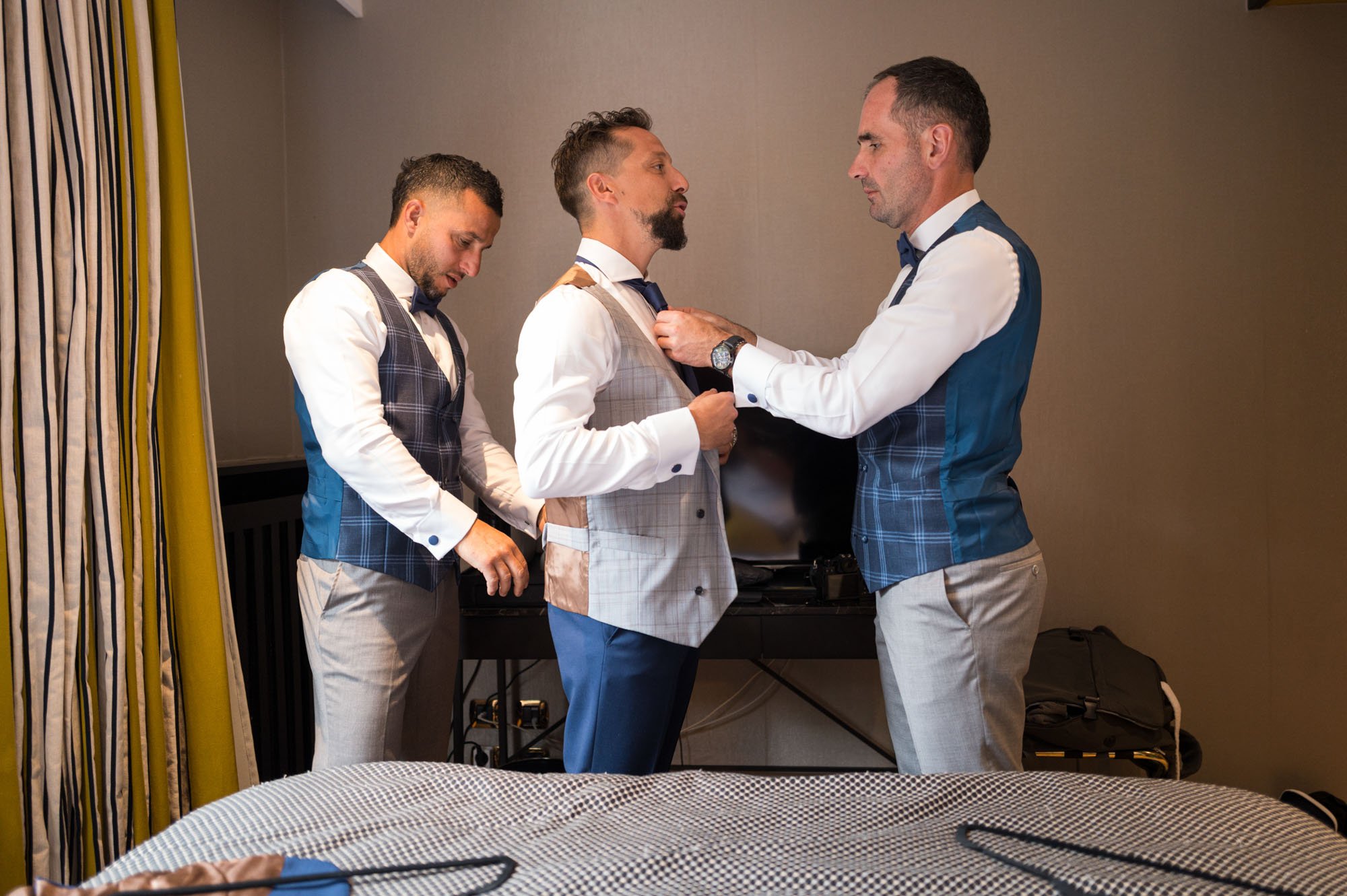 Quatre hommes en costumes se préparent dans une chambre, deux ajustent la cravate de l'homme au centre, un autre ajuste le costume, et le quatrième est concentré sur ses propres vêtements.