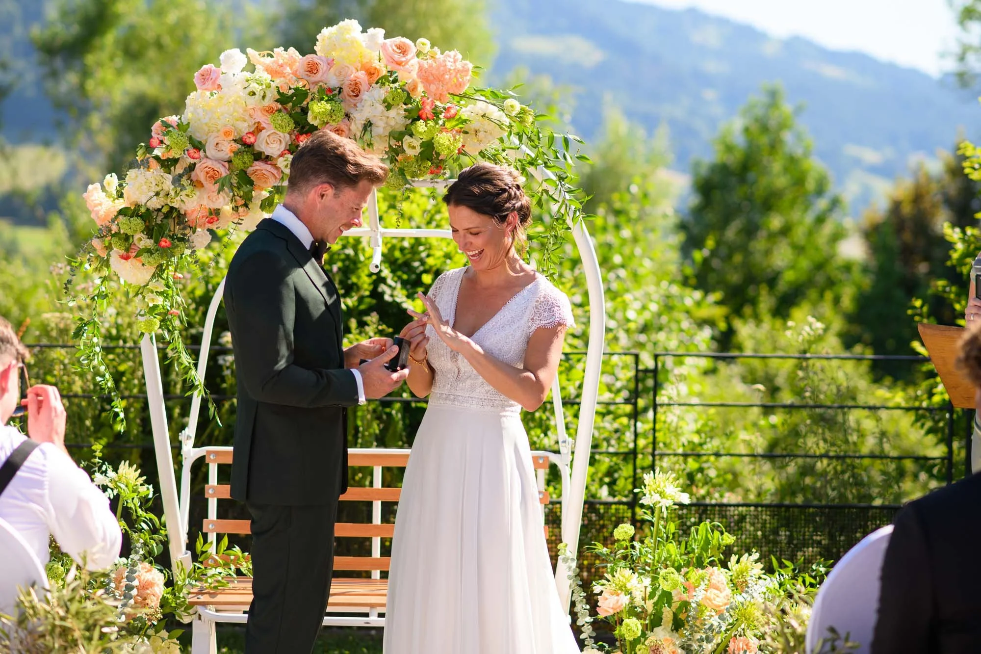 Un couple échange des vœux de mariage en plein air sous un décor floral, avec un fond de montagnes et de verdure, le jour.