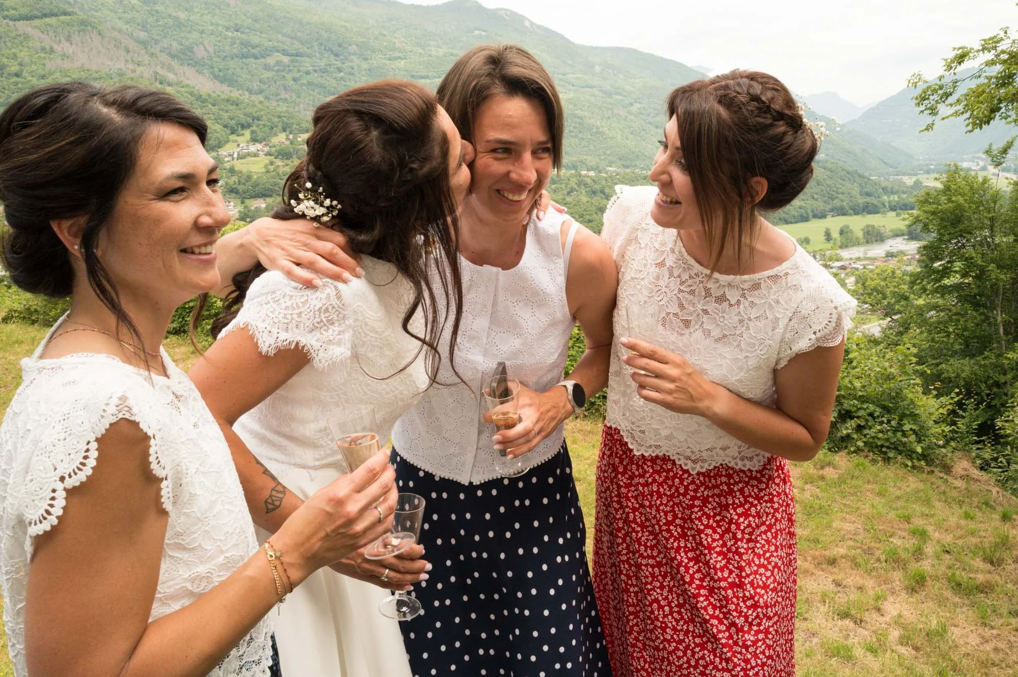 Quatre femmes souriantes en tenues blanches et rouges, se regroupant pour une photo en extérieur avec un paysage de montagne et de verdure en arrière-plan, tenant des flutes de champagne.