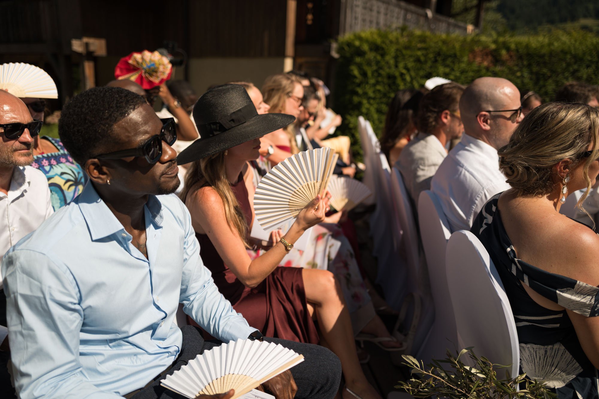 Groupe de personnes assises en plein air sous le soleil lors d'un événement, certaines portent des chapeaux et des éventails, habillées en tenues formelles ou décontractées, avec un mur de buissons en arrière-plan.