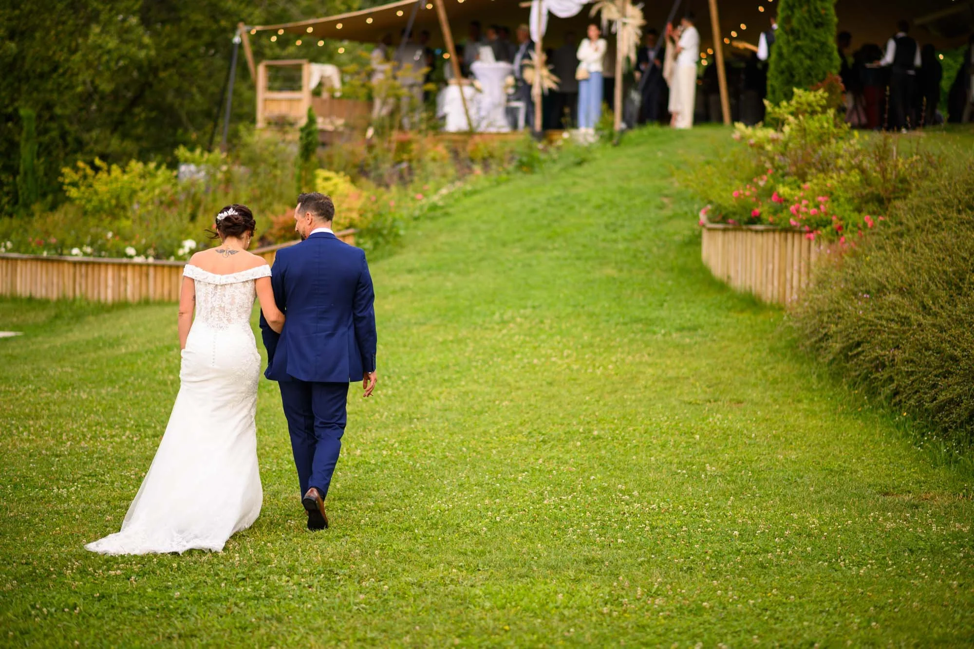 Un couple de mariés marche dans un jardin lors d'un mariage en extérieur, avec un groupe d'invités sous une tente en arrière-plan.
