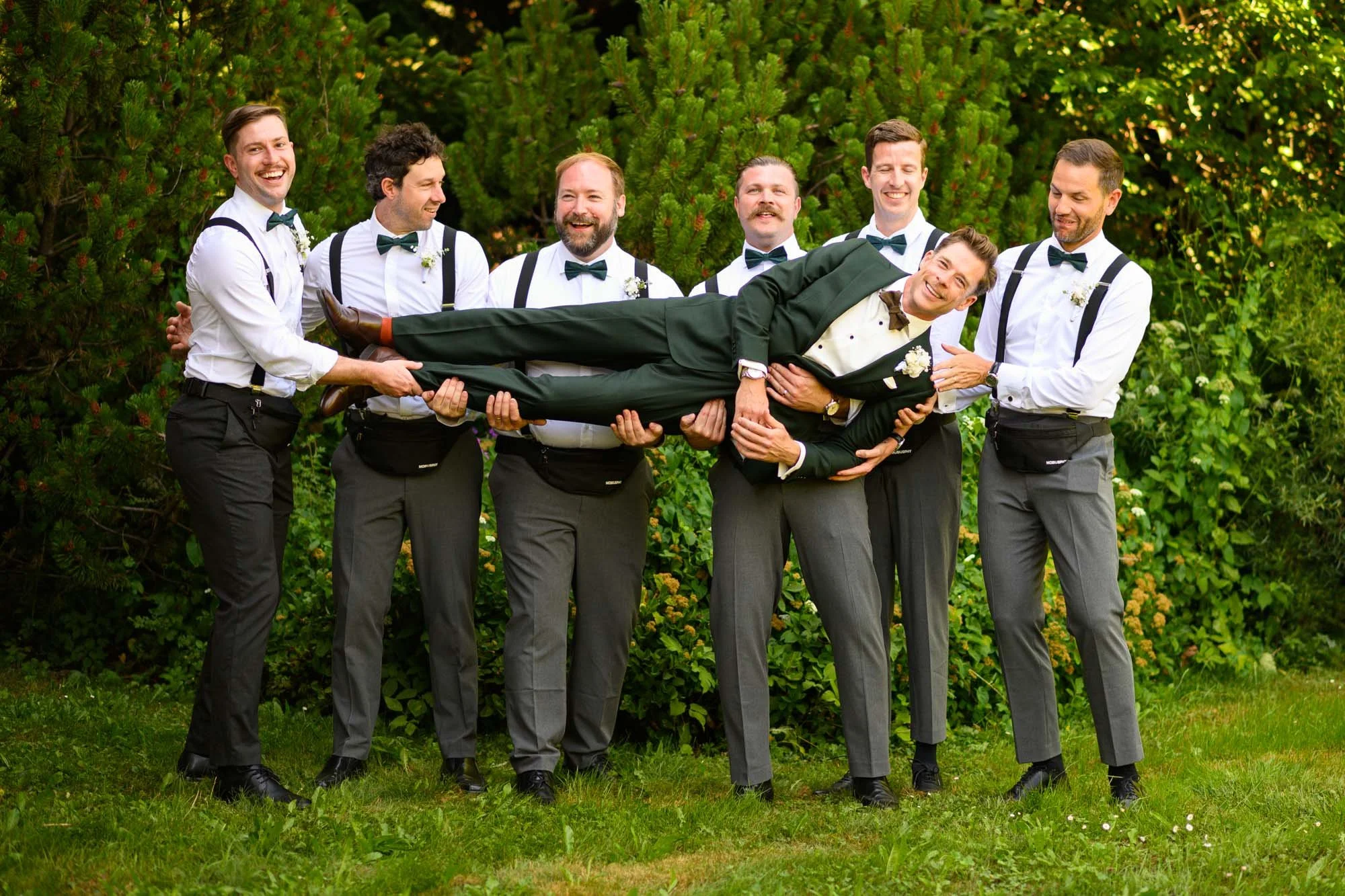 Groupe de sept hommes habillés en costumes gris et chemises blanches, avec des nœuds papillons, tenant un homme portant un costume foncé à l'horizontale, dans un jardin avec des arbres verts