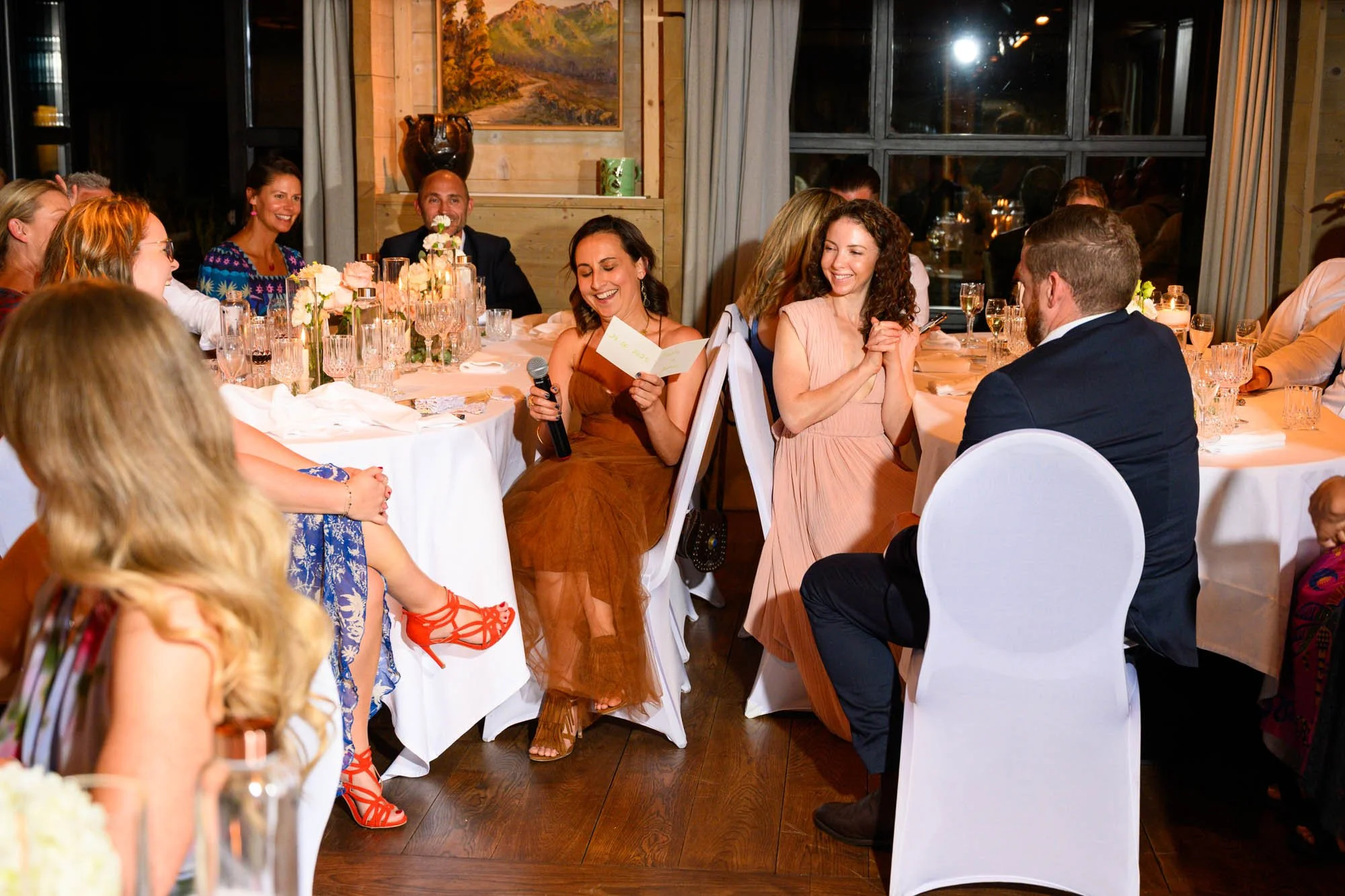 Un groupe de personnes assises autour d'une table lors d'une réception ou d'un dîner, avec deux femmes en conversation et une femme en train de lire un message, dans une ambiance chaleureuse et élégante, avec des décorations florales et un ambiance d