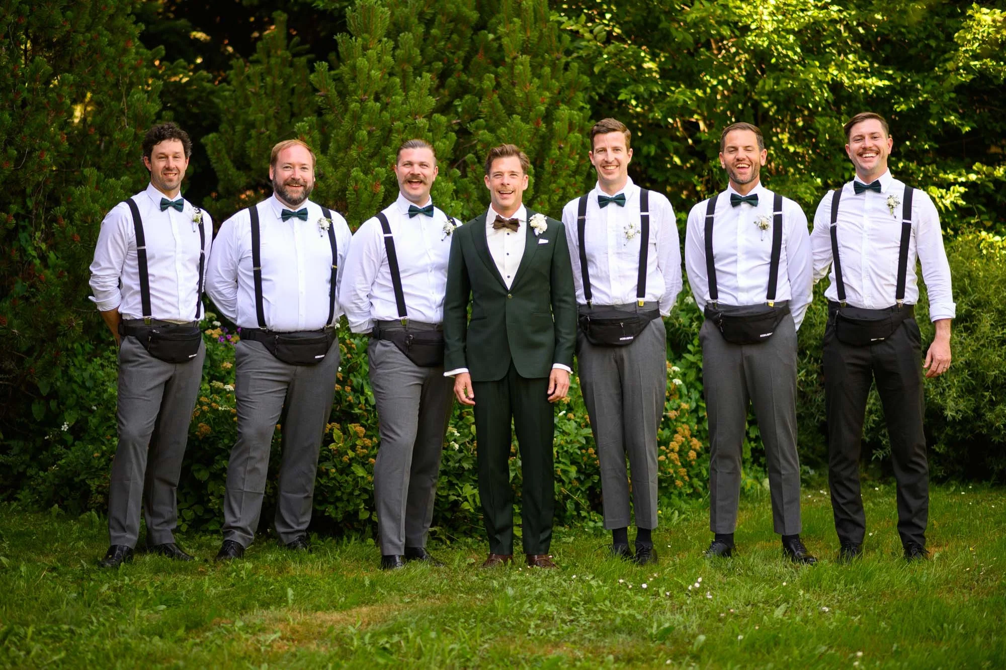 Groupe d'hommes en costume lors d'un mariage en plein air, avec le marié en costume vert foncé au centre, entouré de groomsmen en chemises blanches et pantalons gris ou noirs, dans un jardin avec des arbres verts en arrière-plan.
